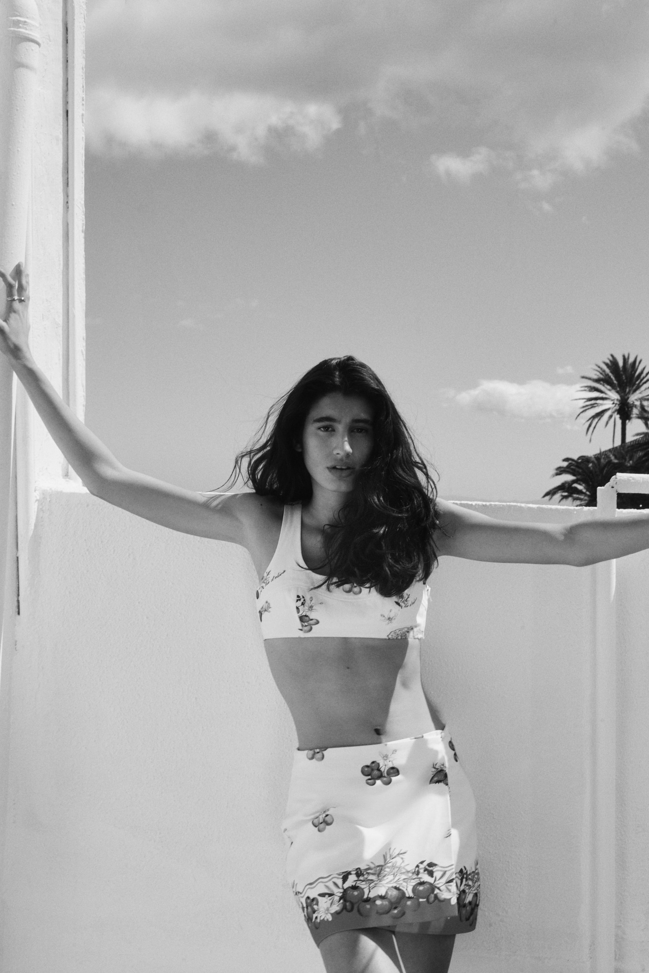 View larger image: A short, fitted A-line skirt is showcased from the front, boasting a wrapover design fastened by a visible covered button on one side. It displays an all-over print of fruits and insects on a light fabric.