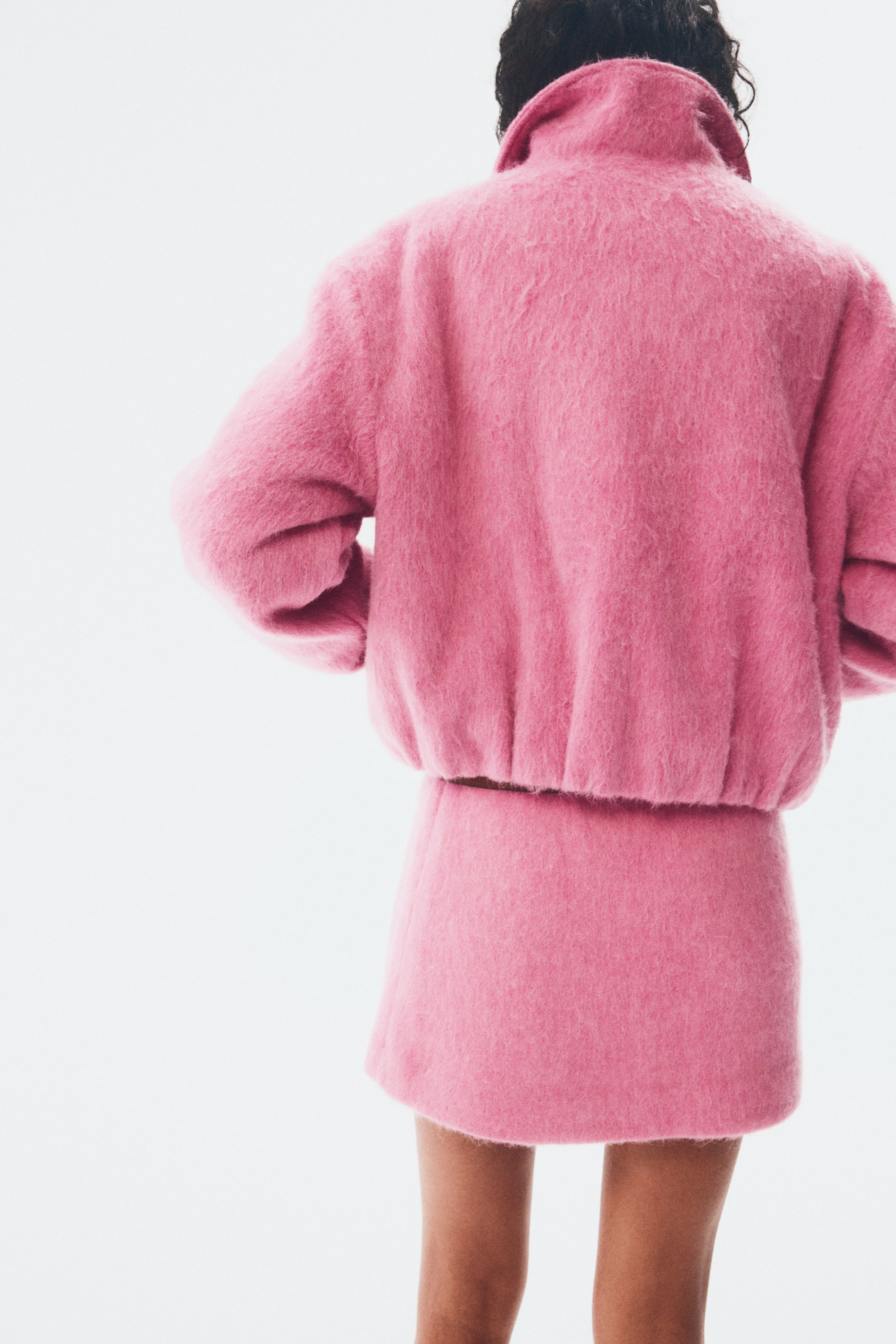 Agrandir l'image: Une veste duveteuse d'un rose vif et une mini-jupe assortie sont présentées de dos. La veste affiche un col montant et un ourlet froncé, tandis que la jupe présente une coupe droite.
