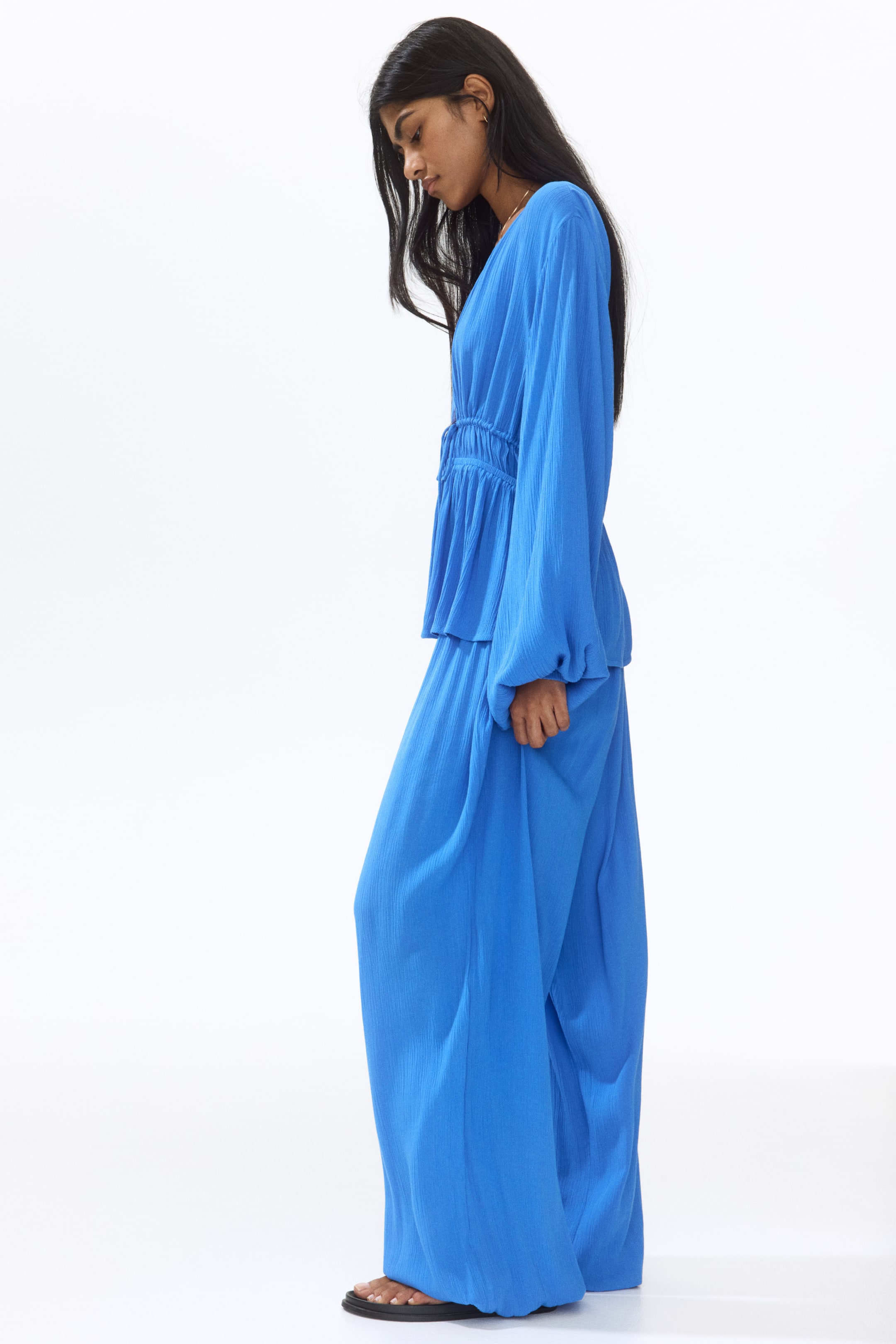View larger image: The model is showcasing blue relaxed-fit trousers crafted in a crinkled viscose weave. These trousers boast balloon legs with covered elastication at the hems, perfectly paired with a matching blue top and sandals.
