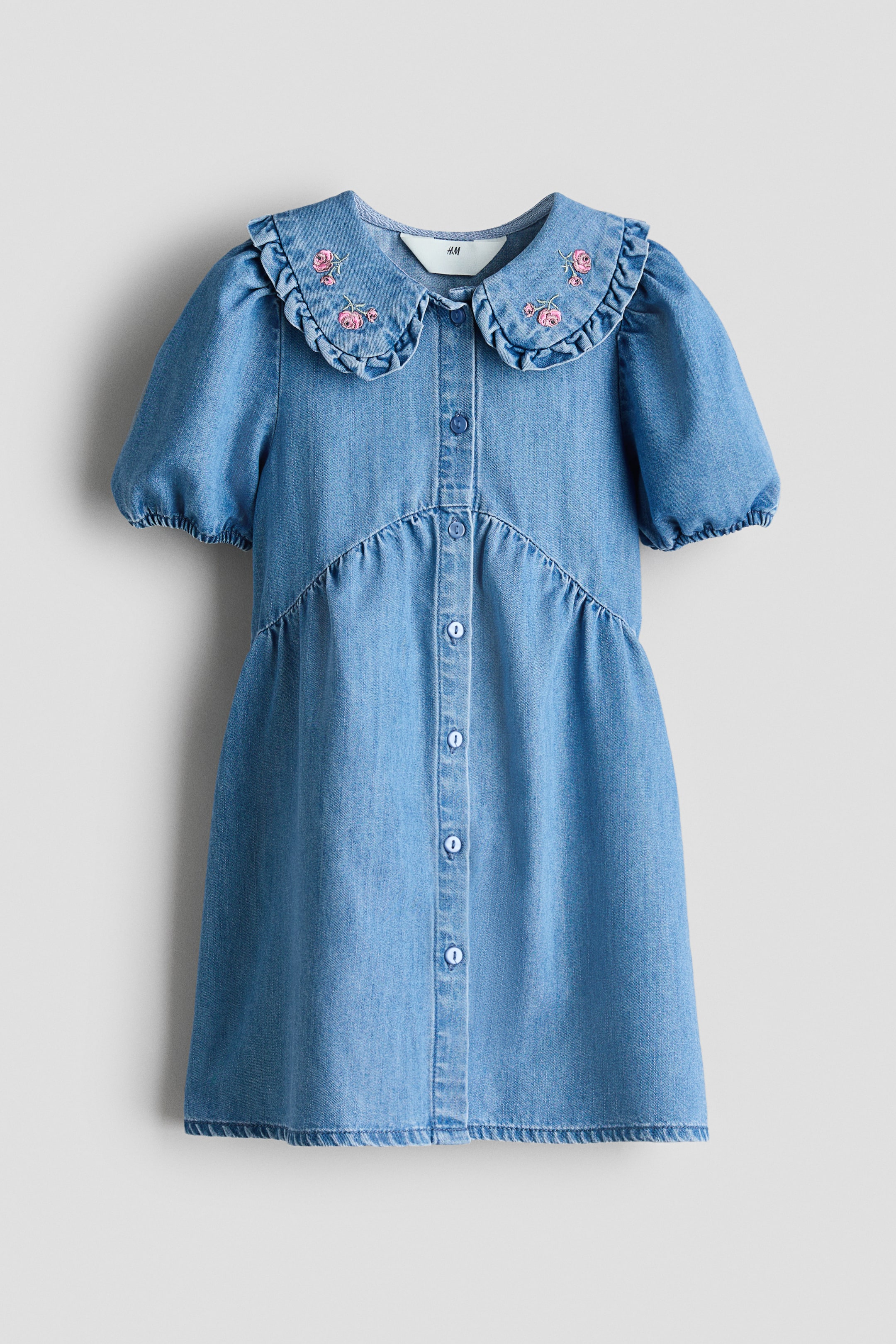 View larger image: A denim dress is shown front-facing, featuring a Peter Pan collar adorned with embroidered pink roses and ruffled edges. It boasts short puffed sleeves, a full-length button placket, and V-shaped seams above a gathered A-line skirt.
