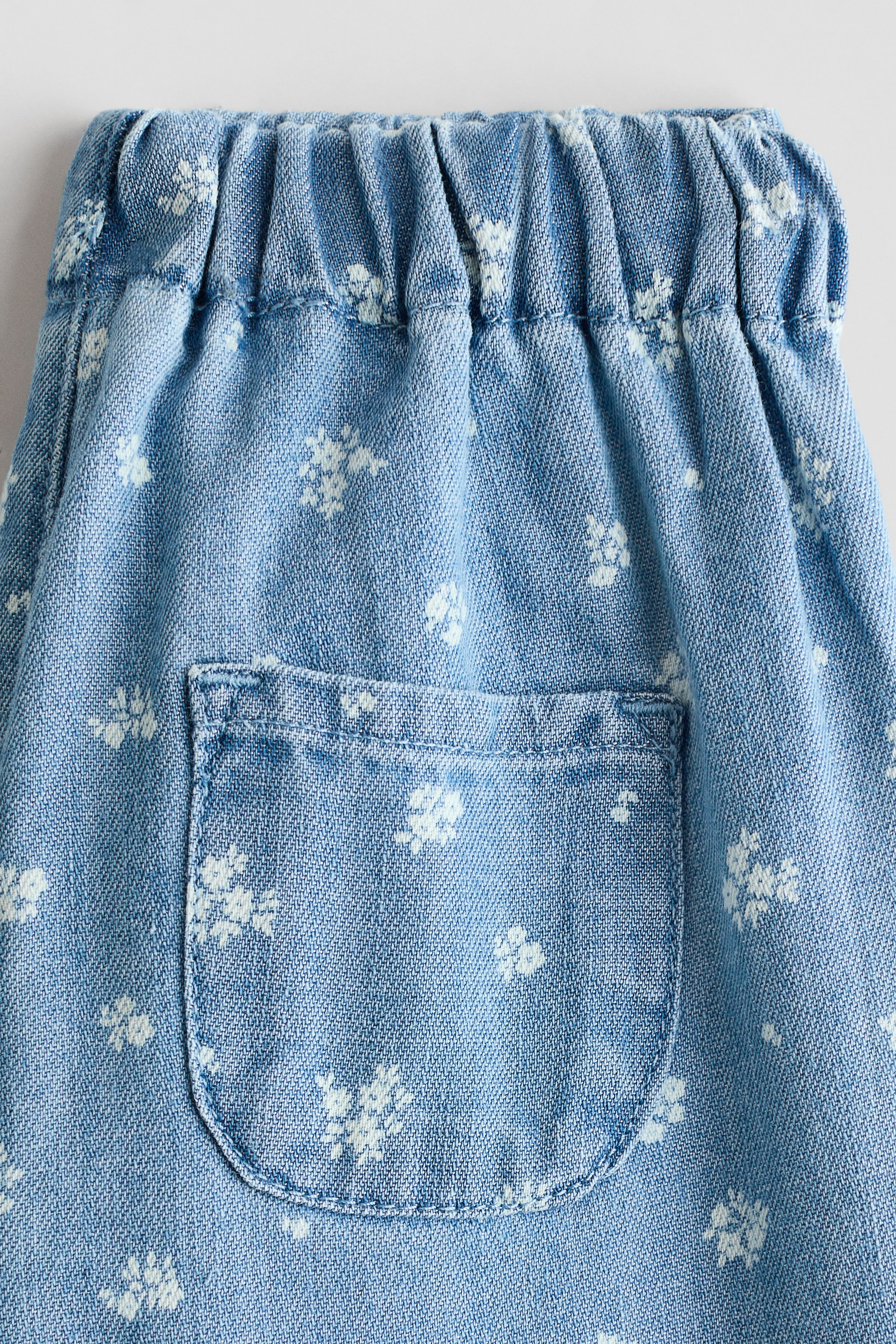 View larger image: A light blue garment features an elasticated, gathered waistband and displays white floral motifs scattered across the fabric. A patch pocket with rounded bottom corners is visible below the waistband.