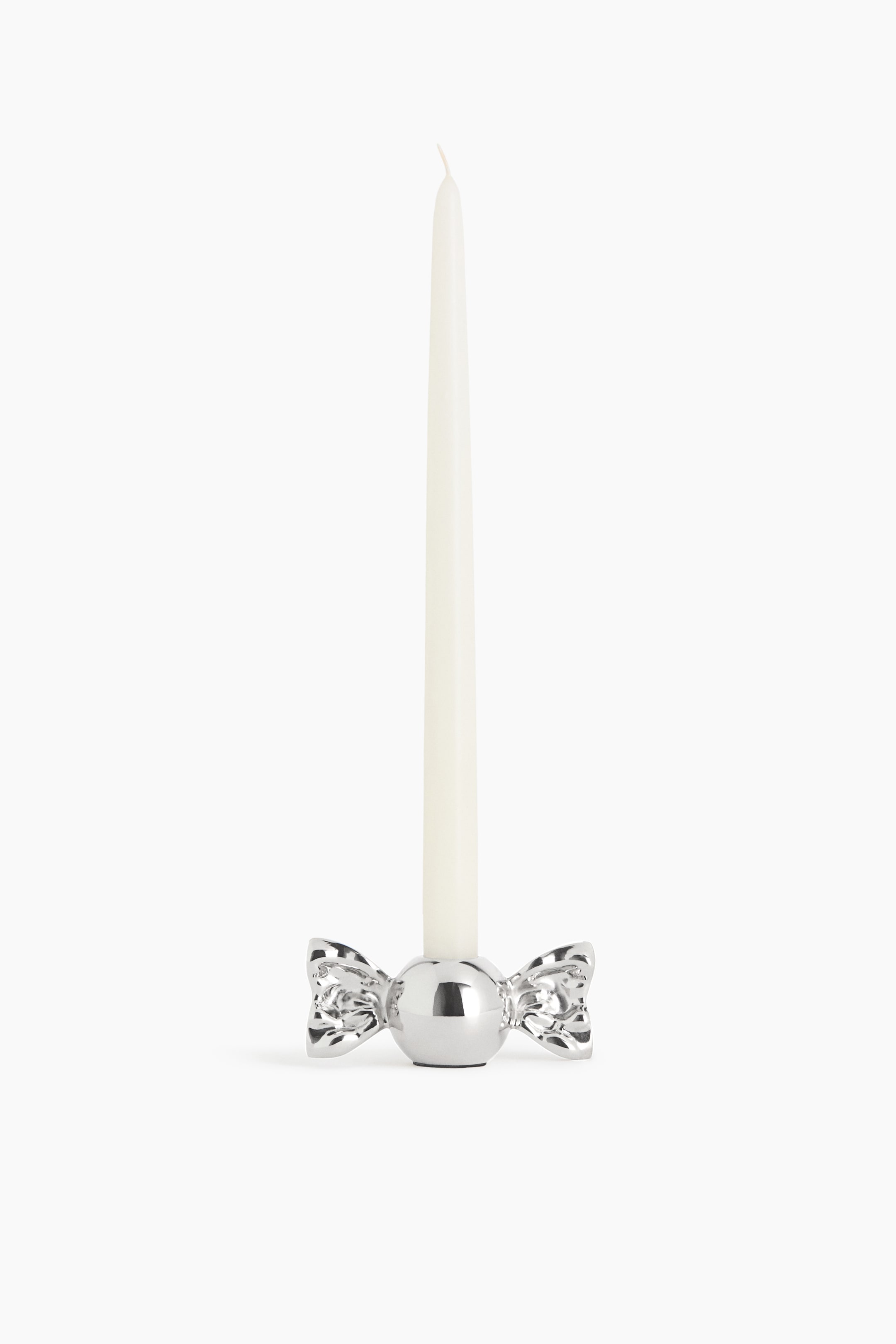 View larger image: A silver metallic candle holder shaped like a wrapped candy is facing front, holding a tall off-white taper candle in its central sphere. The holder features a highly reflective, polished finish with detailed flared wrapper ends extending from both sides.