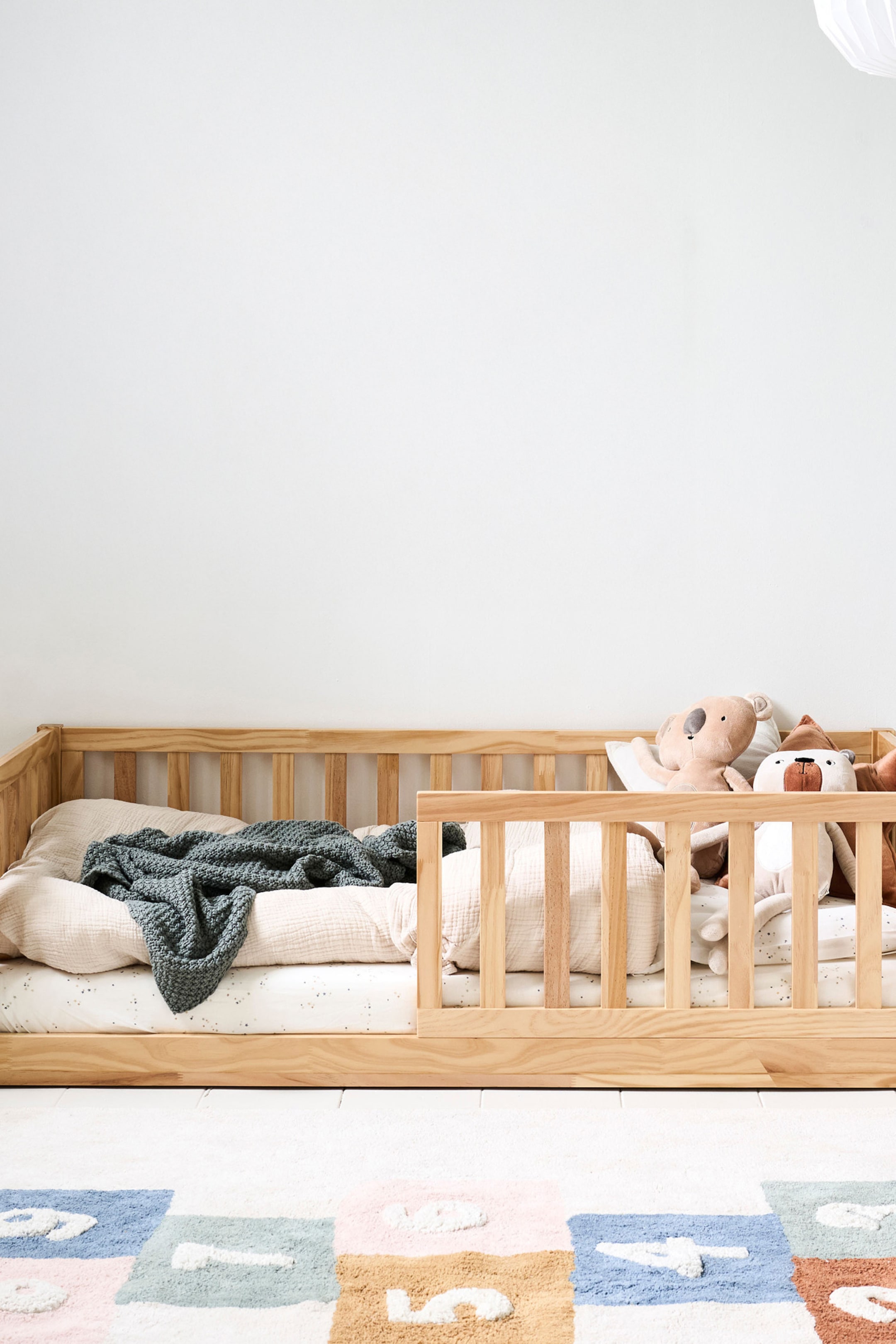 Größeres Bild ansehen: Ein hell getöntes Kinderbett aus Holz präsentiert seinen flachprofiligen Rahmen aus einer frontalen Perspektive. Es verfügt über ein Lattenkopfteil, ein Fußteil und eine lange Seitenleiste mit einem abgesenkten Abschnitt für den Einstieg. Das Bett ist mit einer Matratze, hellfarbener Bettwäsche, einer dunkelgrünen Strickdecke und verschiedenen Stofftieren ausgestattet.