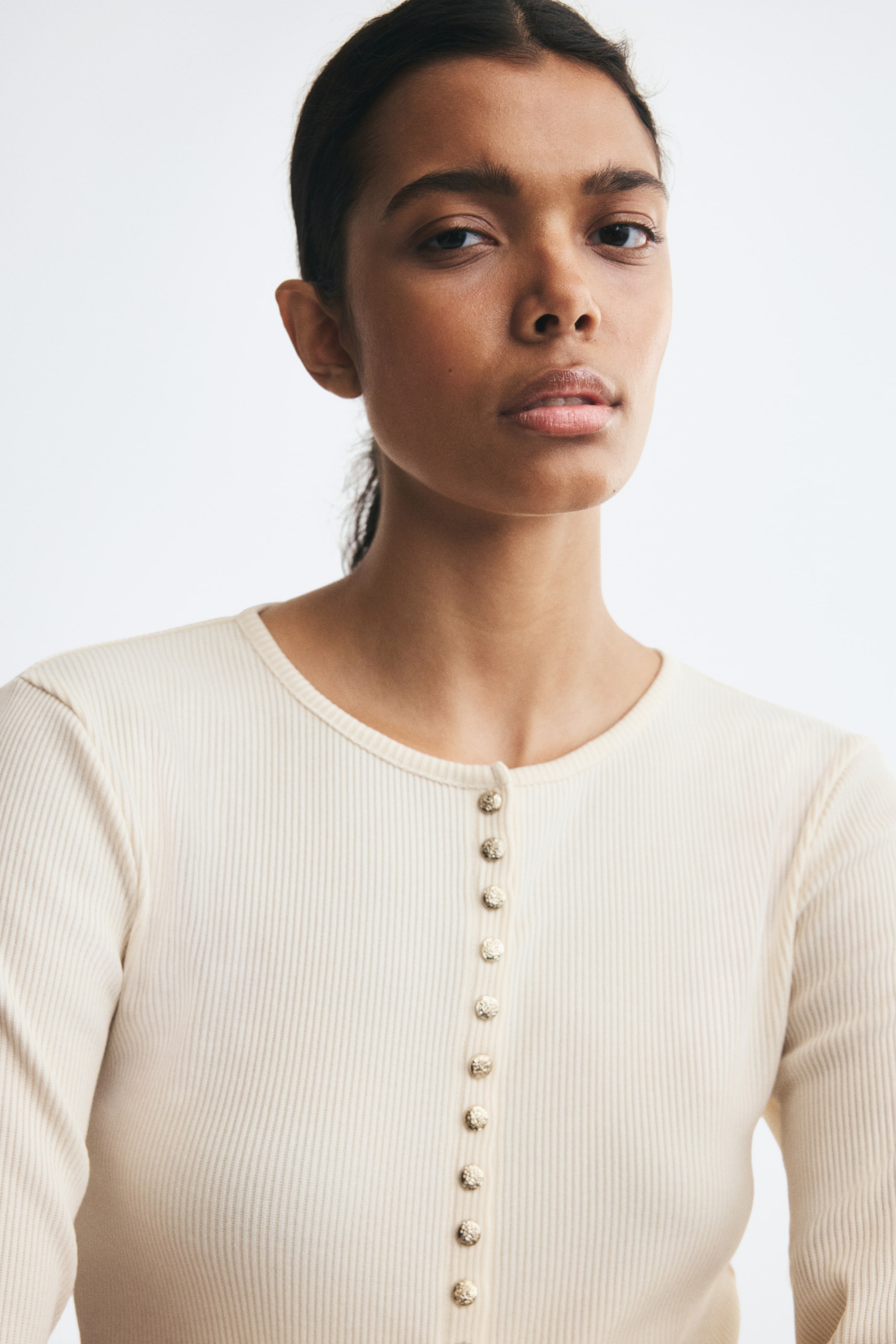 View larger image: A fitted off-white rib-knit cardigan is worn by the model, featuring a round neckline and a row of metallic press studs with a hammered finish down the centre front. The close-up view highlights the fine ribbed texture and detailed buttons of the garment.