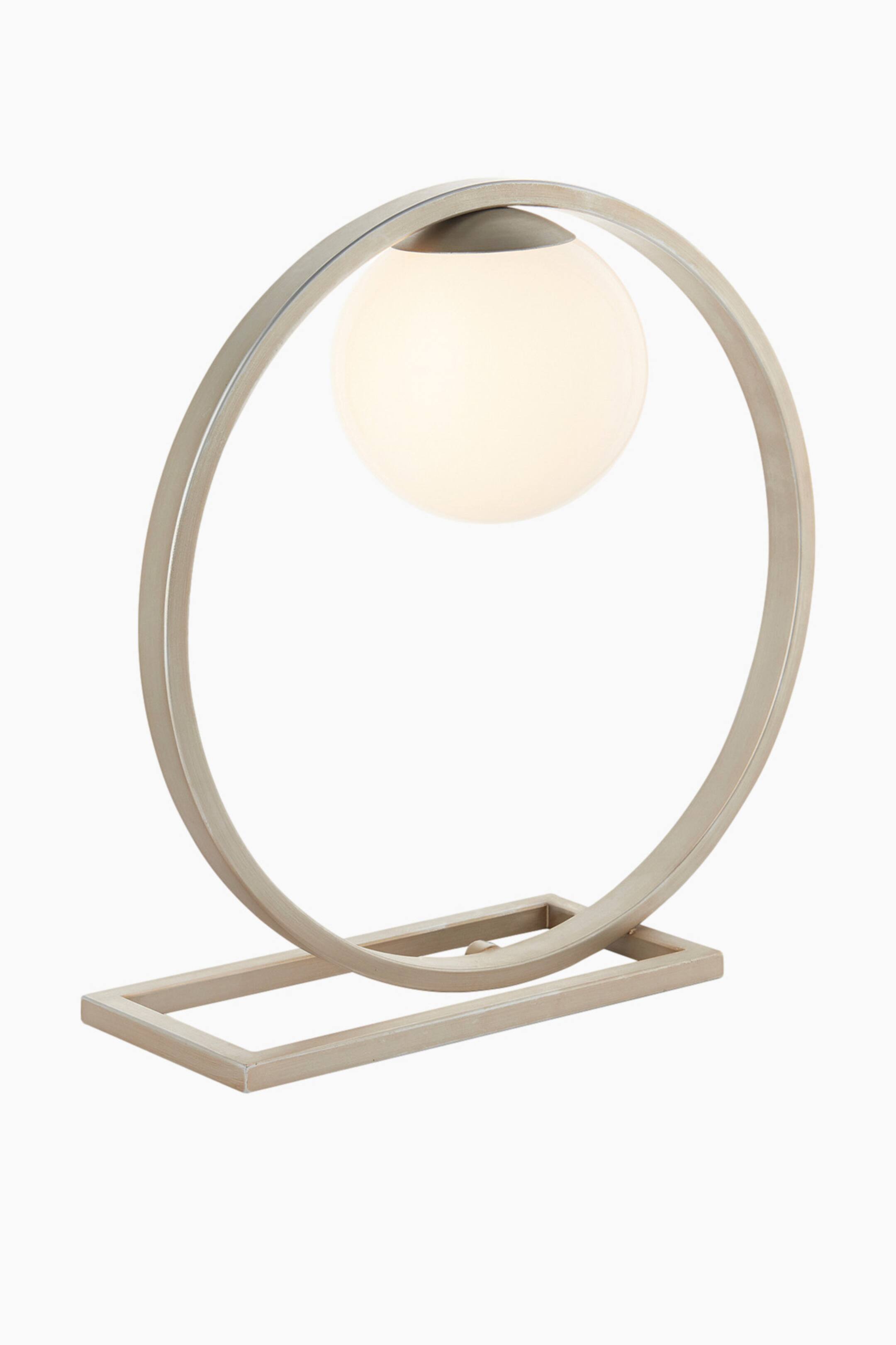 View larger image: A table lamp featuring an open circular, brushed champagne-coloured metal frame that suspends a glowing, frosted white globe light within its upper curve. The frame connects to a rectangular base of the same metal.
