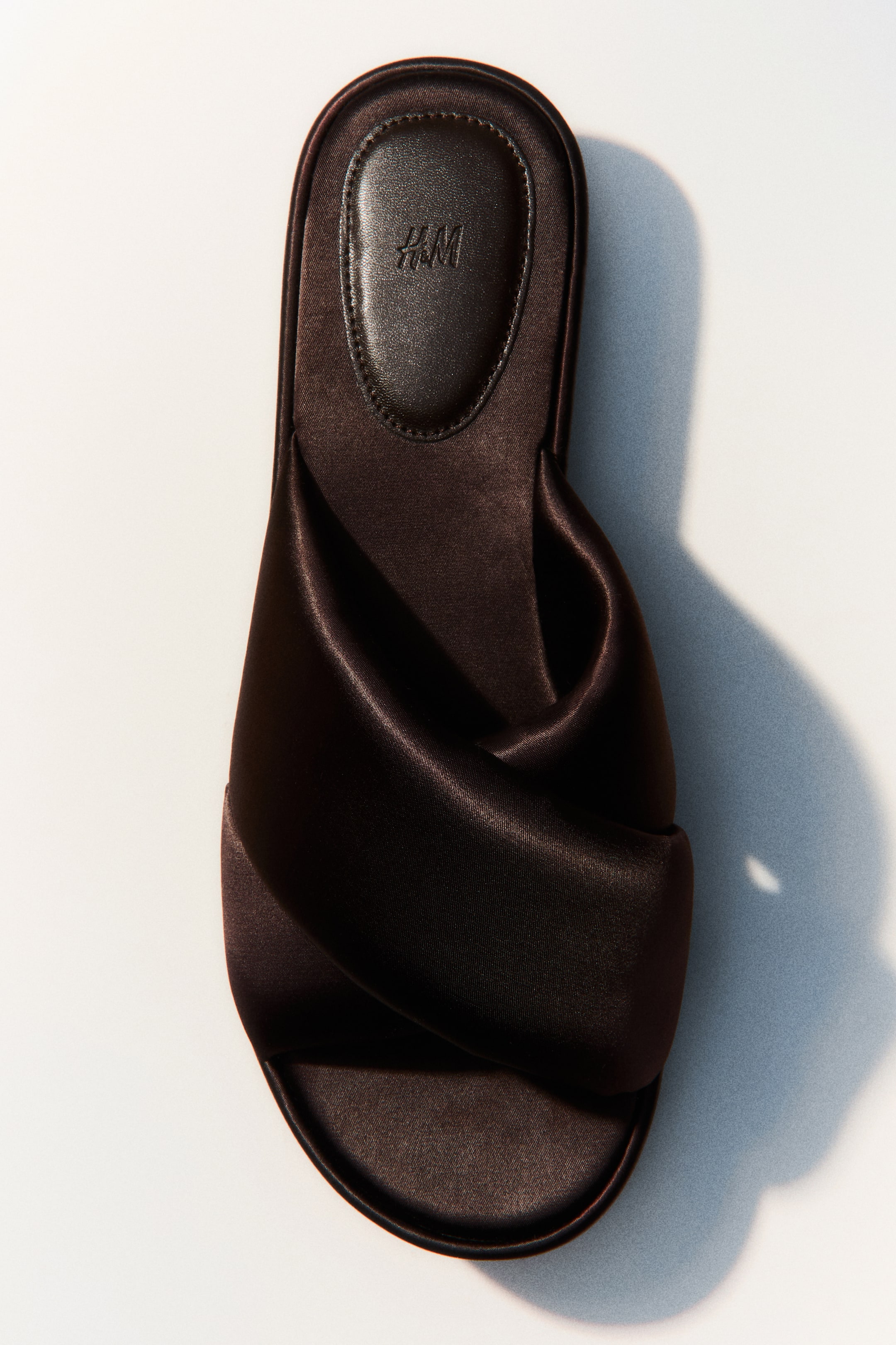 View larger image: A dark brown satin sandal is shown from a top-down perspective. It features a wide, twisted satin strap crossing over the foot and a dark padded insole embossed with the H&M logo.