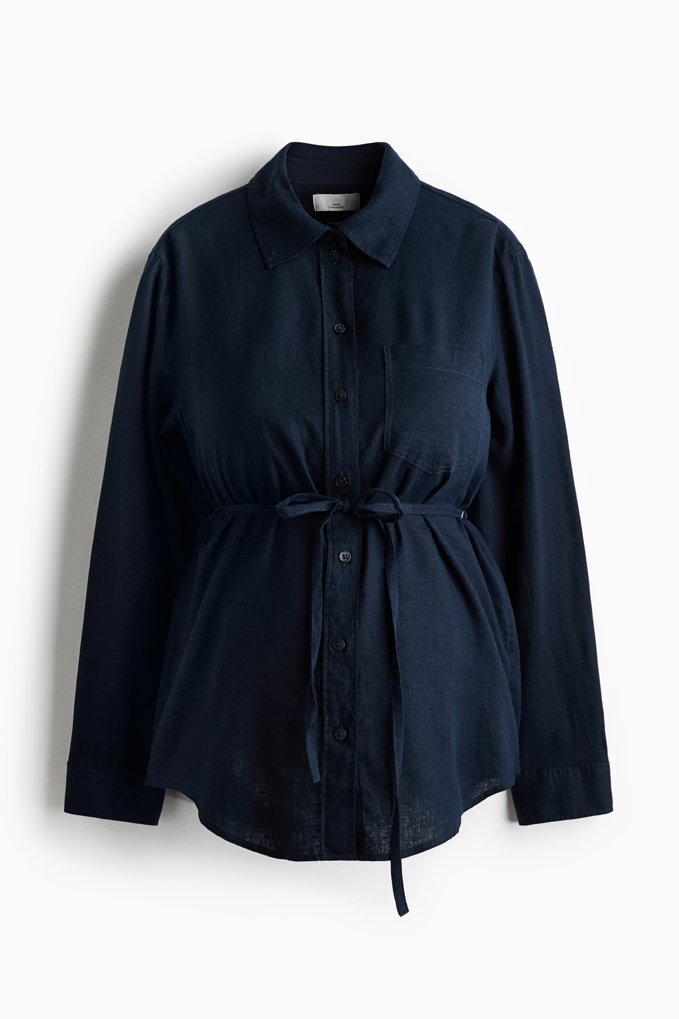 View larger image: A dark blue shirt is showcased from the front, featuring a pointed collar, a button-front placket with dark buttons, and long sleeves with buttoned cuffs. A self-fabric tie cinches the waist, forming a bow at the front and creating gathers, with a single patch pocket visible on the left chest and a curved hem.