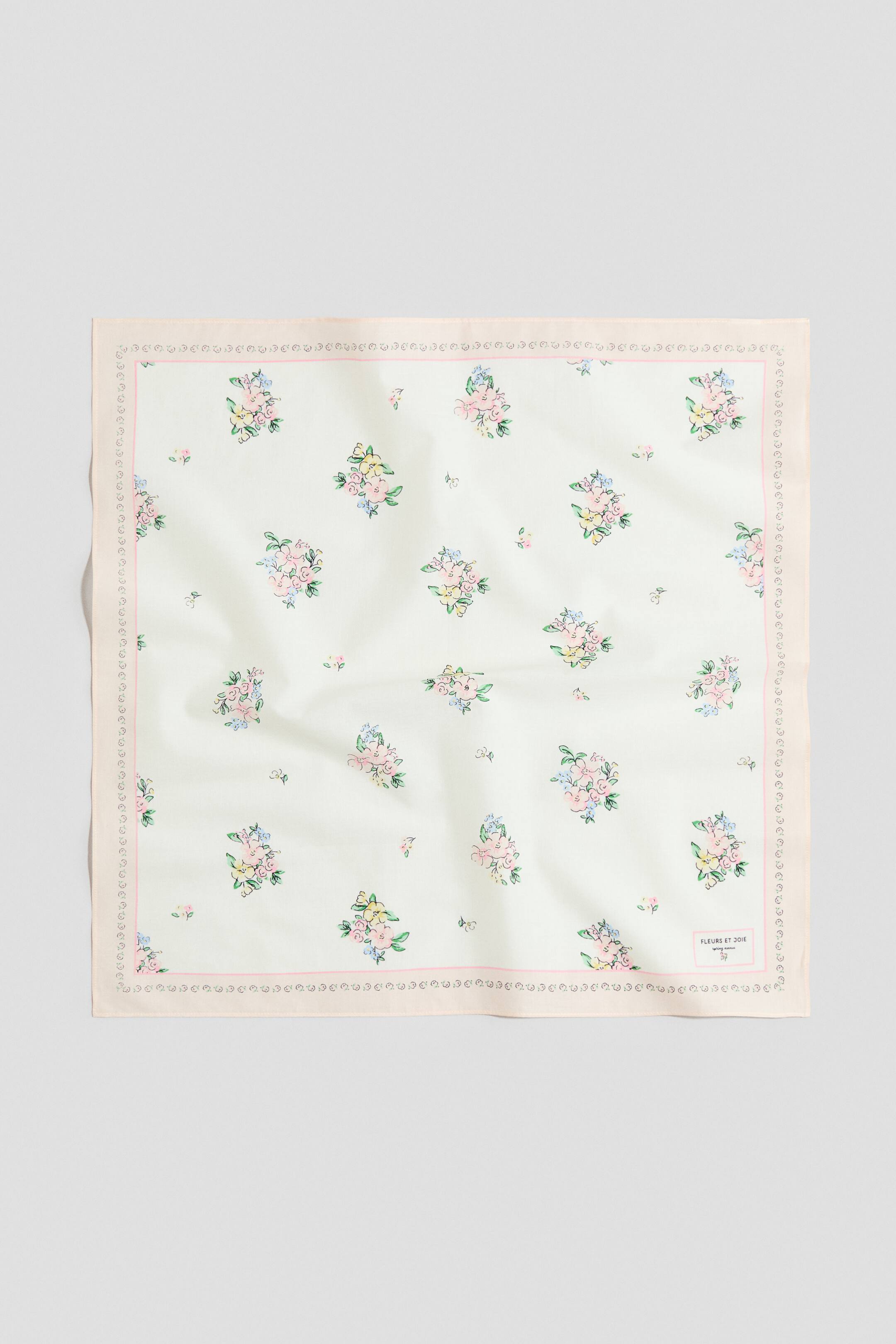 Ampliar la imagen: Un pañuelo cuadrado está extendido, mostrando un fondo blanco con un estampado floral disperso de flores rosas, azul claro y amarillas, y hojas verdes. Presenta un borde de color melocotón claro con un patrón decorativo interior y una pequeña etiqueta "FLEURS ET JOIE" en la esquina inferior derecha.