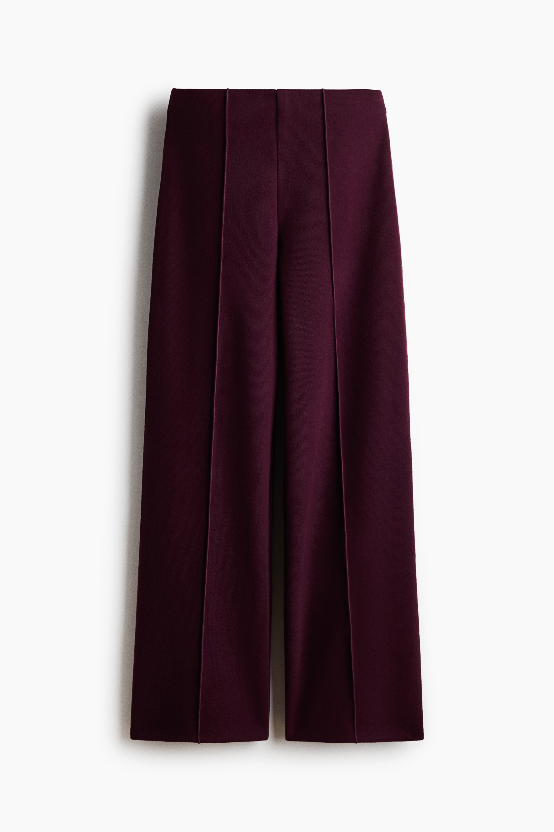 Pantalones de punto de corte holgado en color ciruela intenso, con cintura elástica y marcadas pinzas cosidas al frente de las piernas rectas, que lleva puestos la modelo. Los pantalones se combinan con una camiseta de manga corta a rayas y zapatos planos de color claro.
