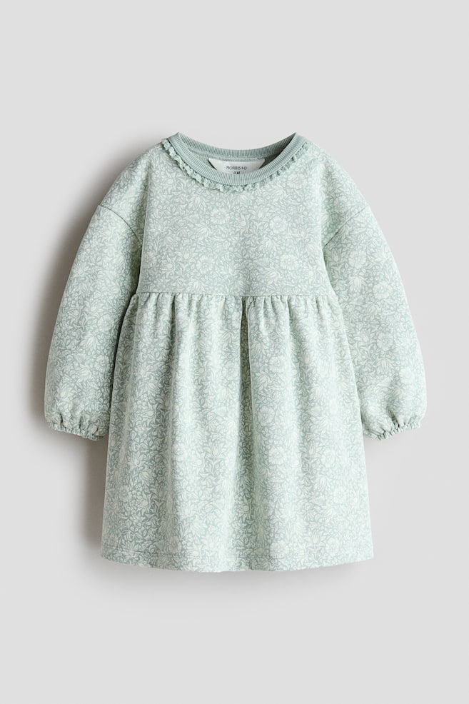 A light green long-sleeved dress is shown from the front, featuring an all-over white floral print. The dress is designed with a round, rib-trimmed neckline displaying a delicate lace trim, dropped shoulders, voluminous sleeves with elastic cuffs, and a gathered waist flowing into a flared skirt. - 1