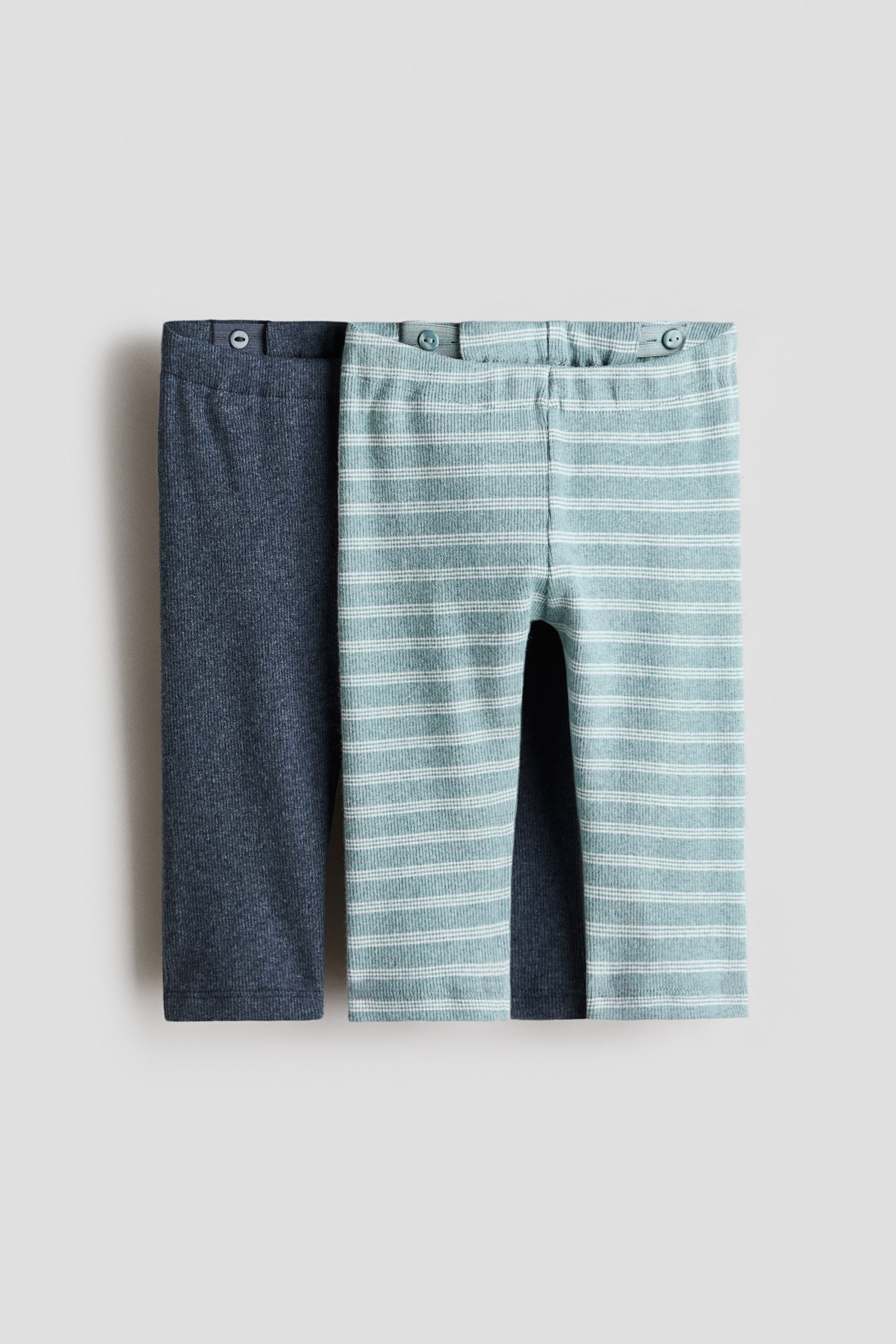 Visa större bild: Två par H&M Adorables leggings ligger platt, vända framåt, och visar upp en ribbad jerseystruktur. Ett par är i en solid mörkblå-grå färg, medan det andra är ljusblå-teal med tunna vita horisontella ränder, och båda har en elastisk midja med synliga interna knappar för justering.