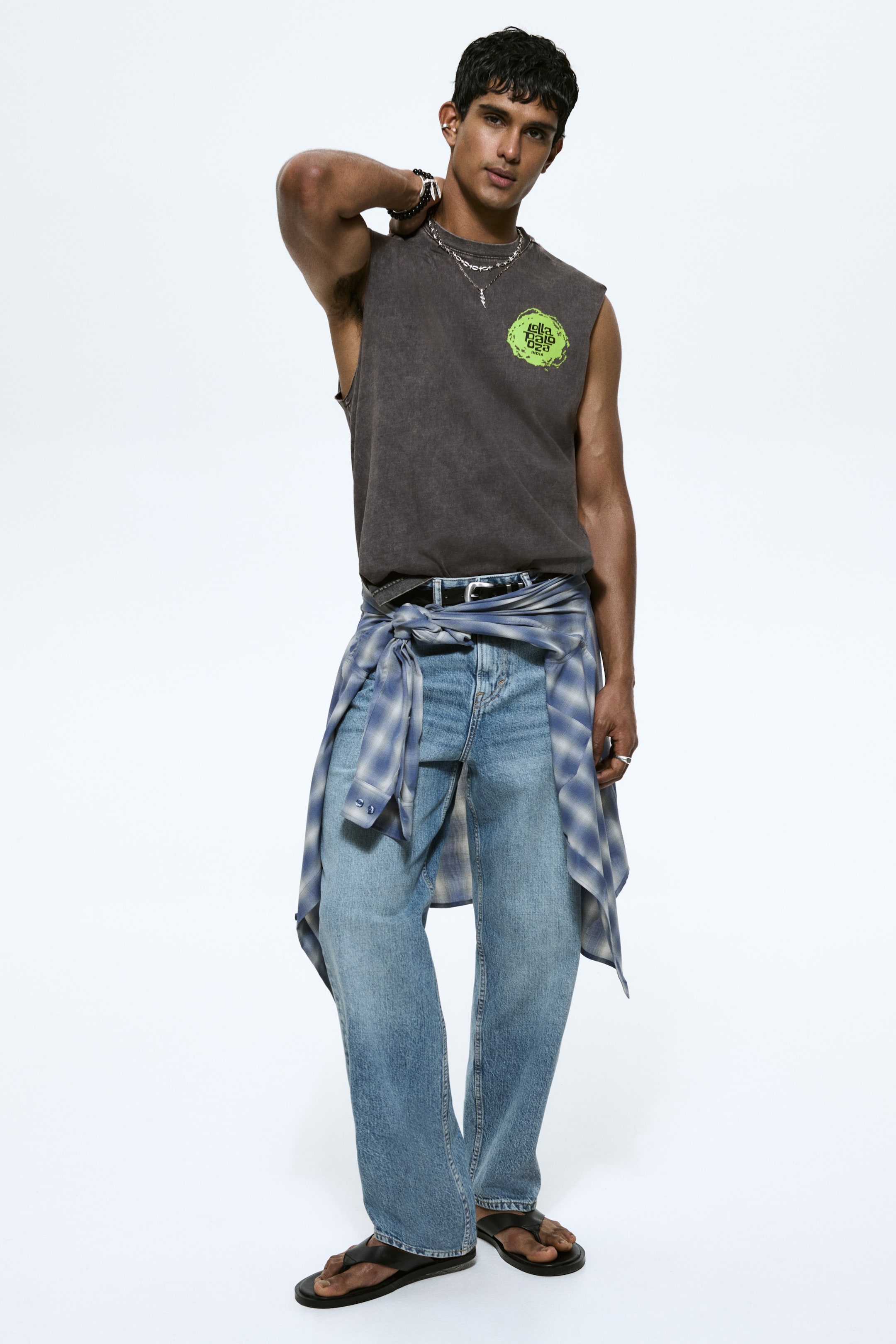 View larger image: A dark grey washed sleeveless T-shirt featuring a neon green graphic on the left chest. Styled with light blue relaxed-fit denim jeans, a black belt, and a blue-and-white checked shirt tied around the waist, with black flip-flops completing the look.