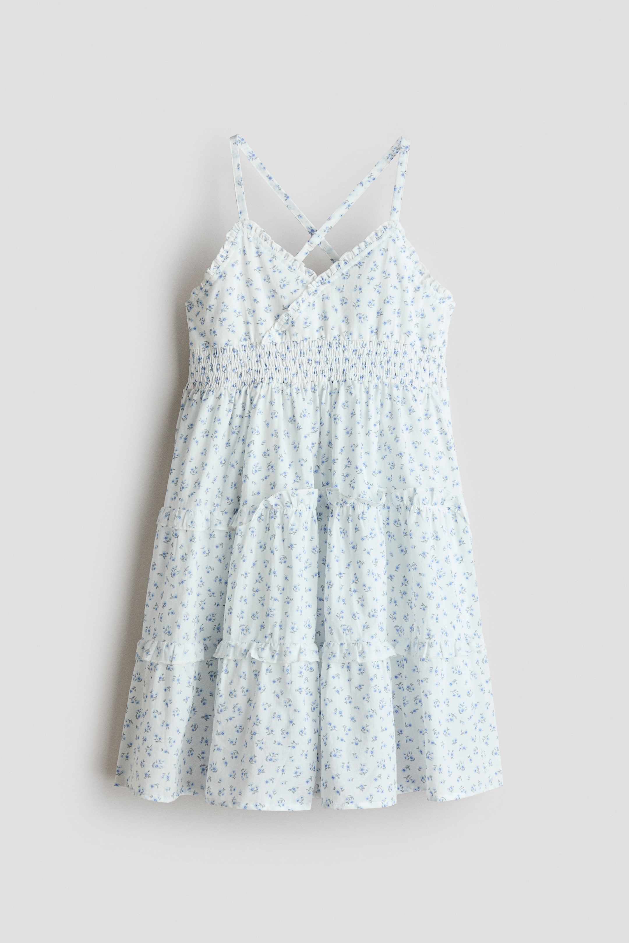 View larger image: The dress is white with a petite blue floral motif, showcased from the front. It features slender criss-cross straps, a shirred elasticated bodice, and a tiered skirt with ruffled edges.