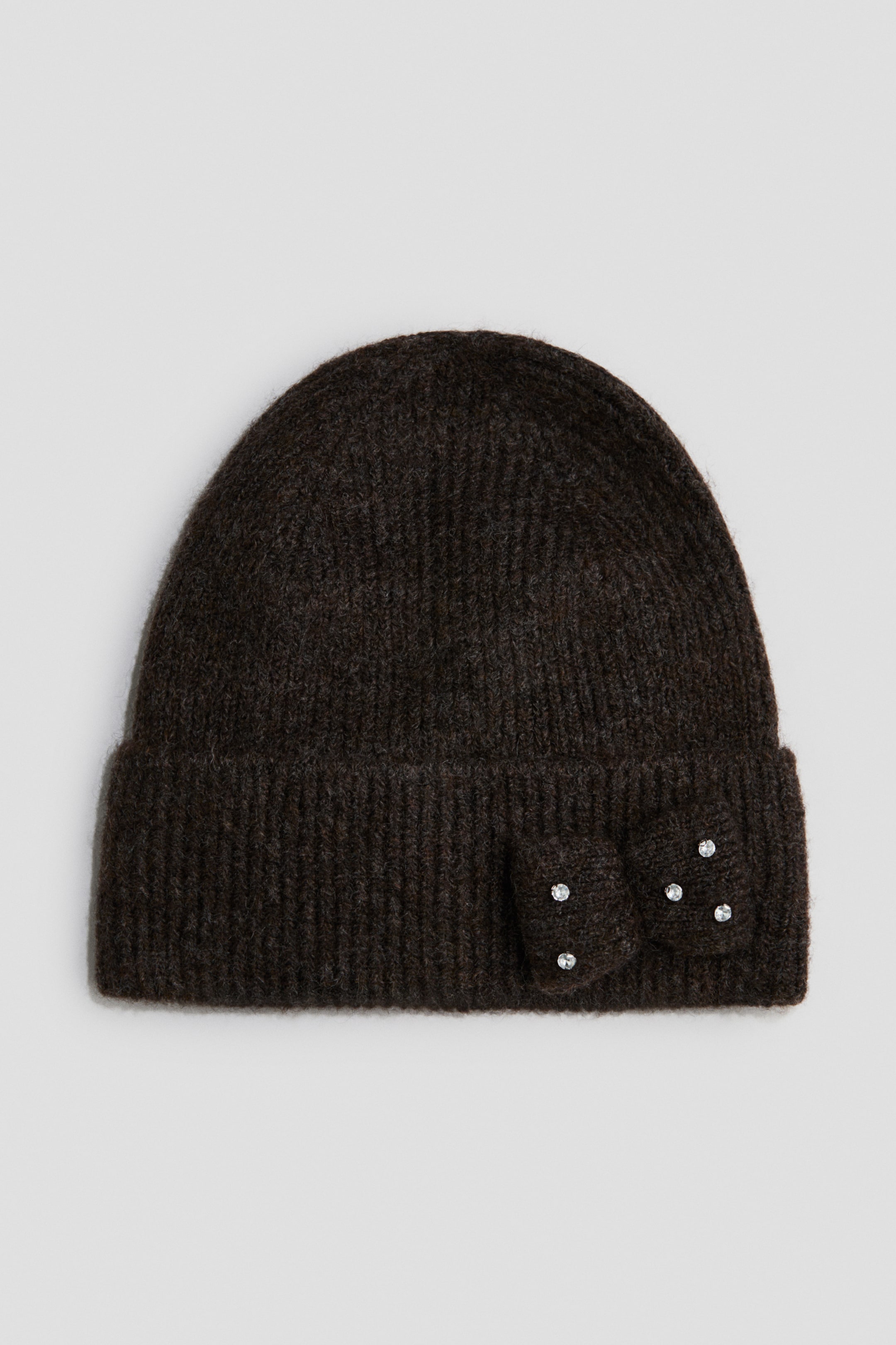 Se større billede: En mørkebrun ribstrikket beanie er vist forfra og har en opfoldet kant. En matchende strikket sløjfe dekoreret med fem små rhinsten er fastgjort på højre side af kanten.