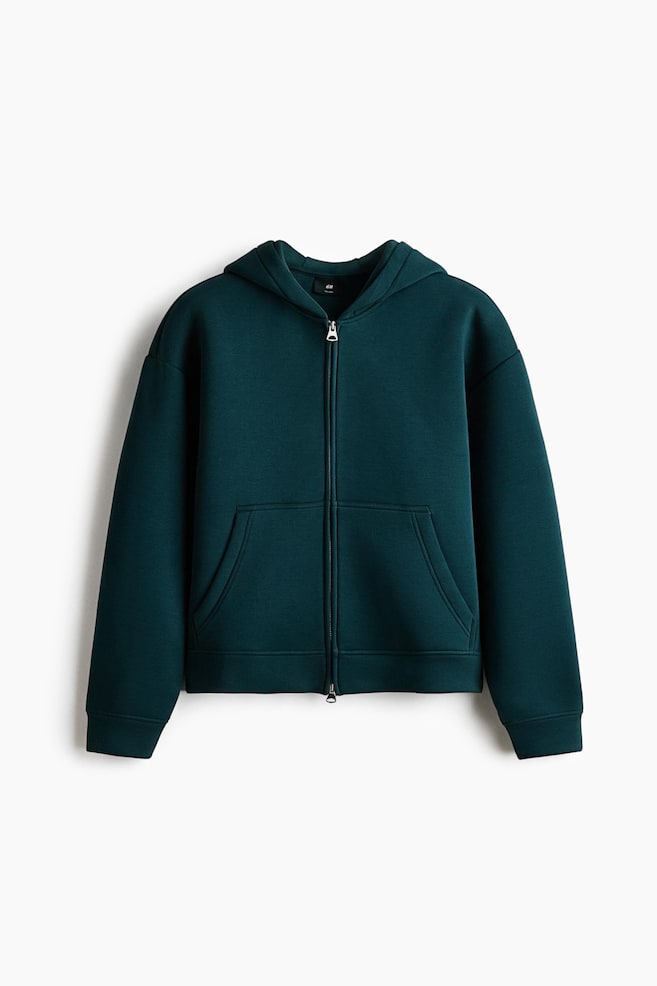 A dark teal, boxy-style zip-up hoodie in a smooth scuba fabric is shown from the front, displaying a loose fit with dropped shoulders and a lined hood. The garment features a full-length front zipper, open split-front pockets, and ribbed cuffs and hem. - 1