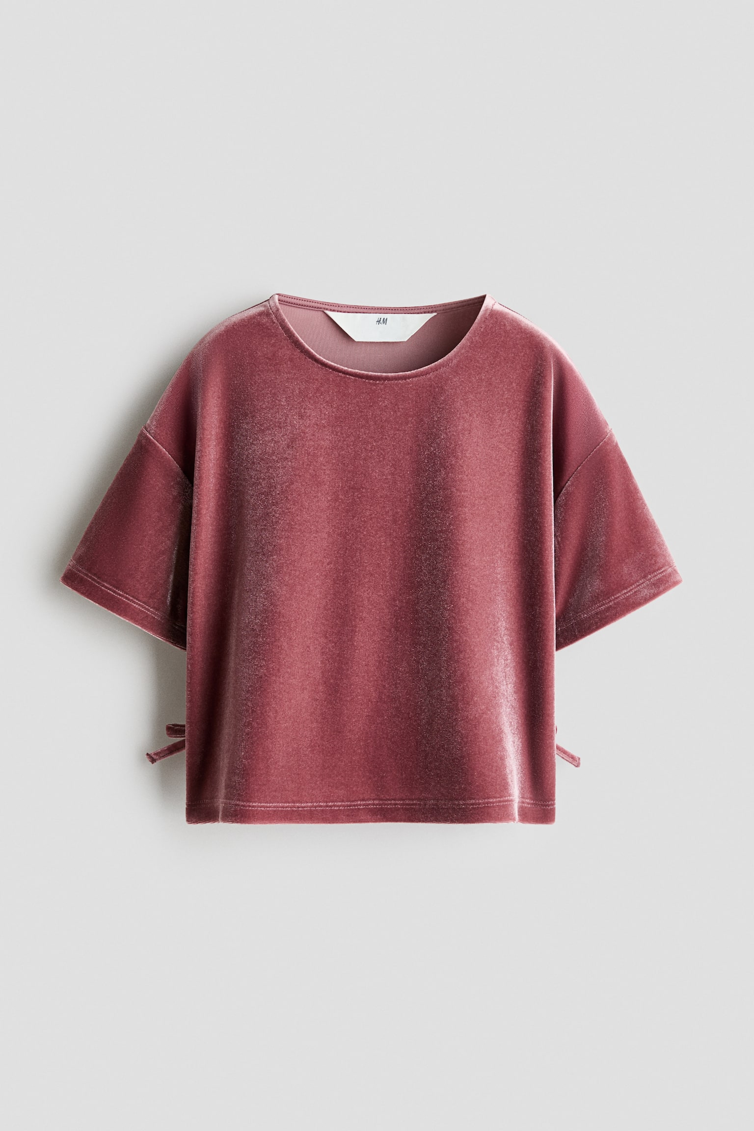 Un t-shirt en velours rose poudrÃ© est prÃ©sentÃ© de face, avec un col rond, des Ã©paules tombantes et de larges manches courtes. Le haut affiche une texture moelleuse et lÃ©gÃ¨rement rÃ©flÃ©chissante, et prÃ©sente de petits dÃ©tails dÃ©coratifs Ã  nouer sur les cÃ´tÃ©s prÃ¨s de lâourlet droit.