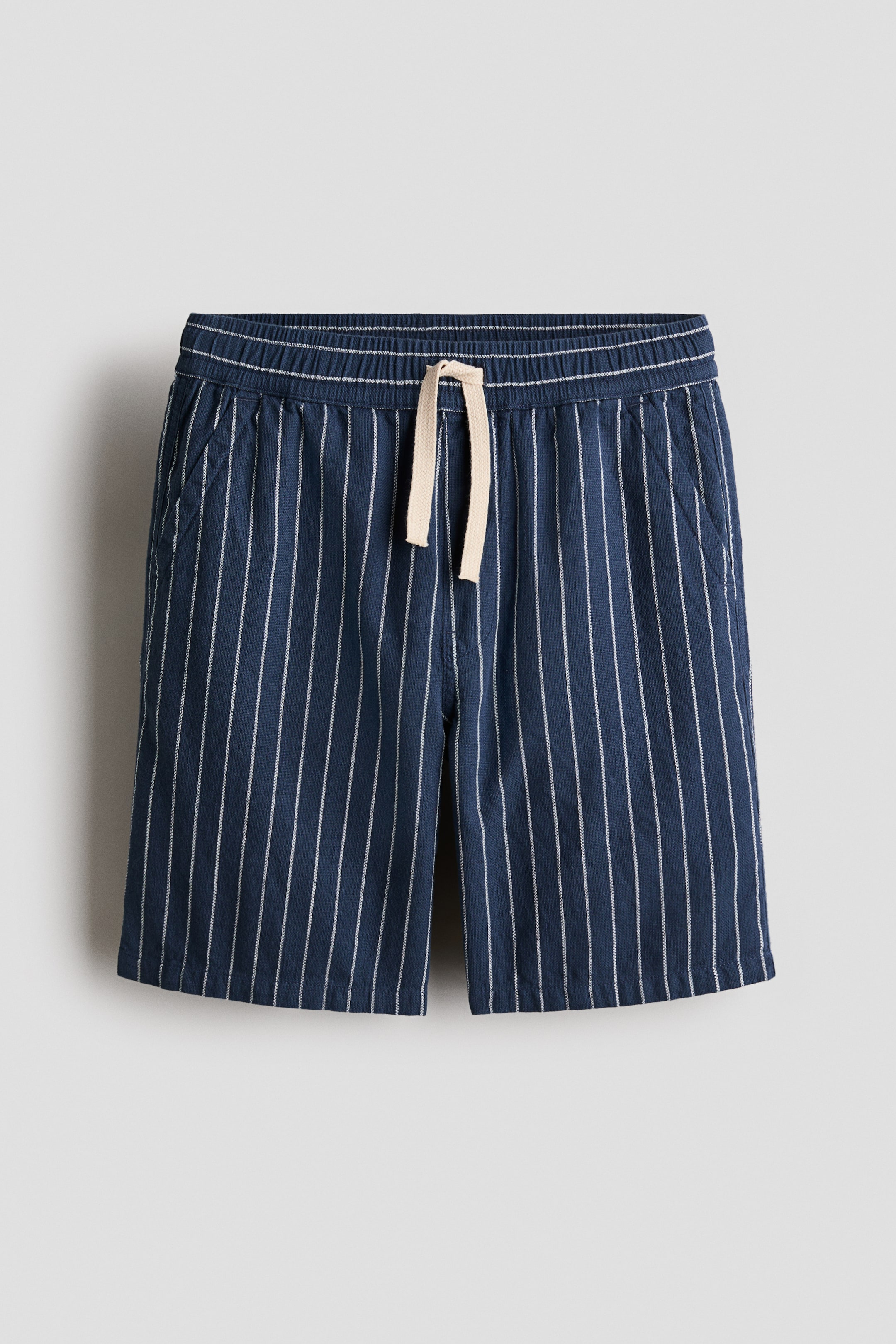 View larger image: Loose-fit shorts showcase a navy blue textured fabric adorned with white vertical pinstripes, front-facing. They come with an elasticated waistband and a light beige drawstring, diagonal front pockets, and a stitched faux fly detail.