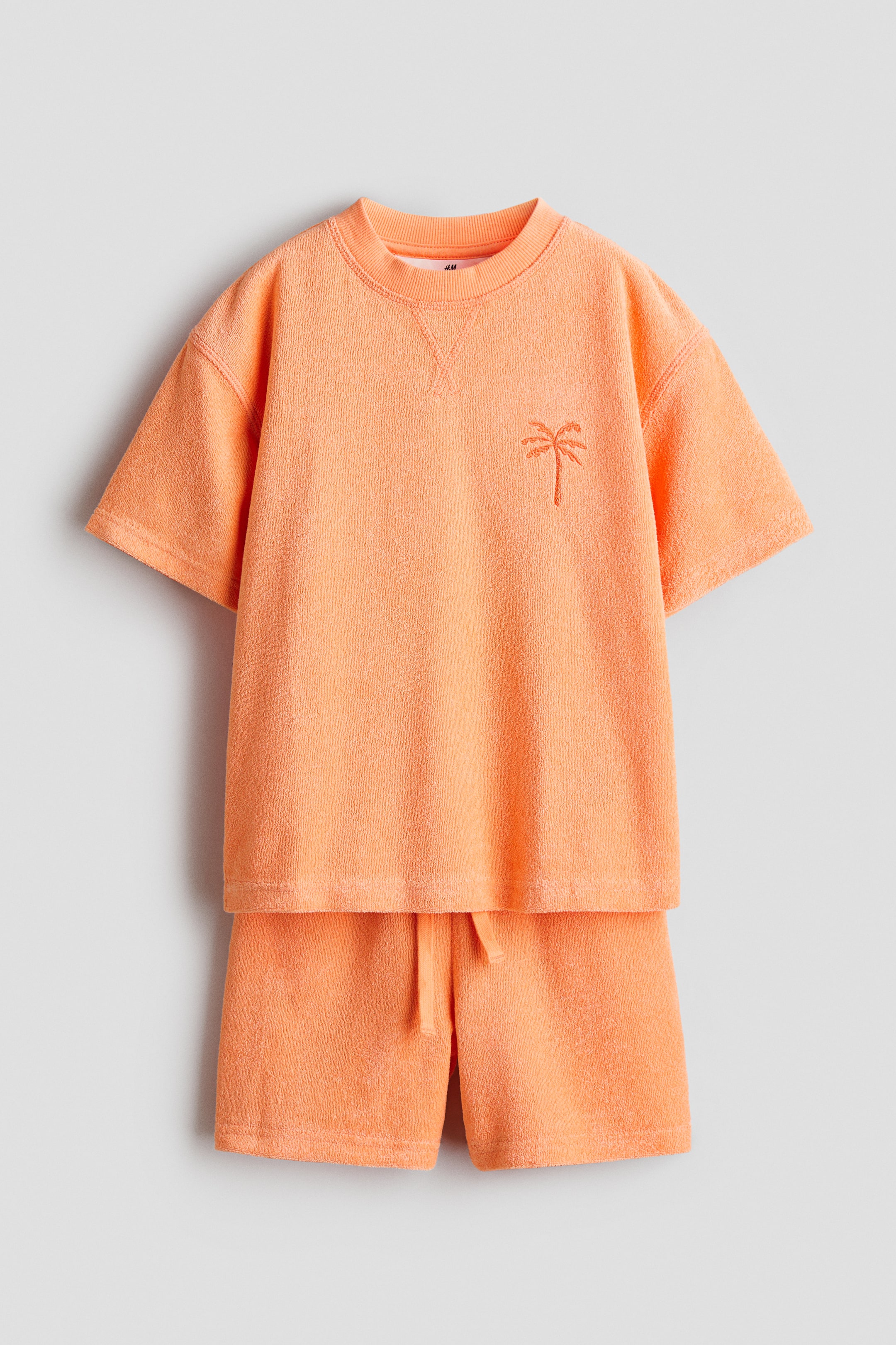 View larger image: The orange terry cloth two-piece set is showcased from the front, consisting of a short-sleeved top paired with matching shorts. The top features a ribbed crew neck with a V-stitch detail and an embroidered palm tree on the left chest. The shorts come with an elasticated waistband complete with a drawstring.