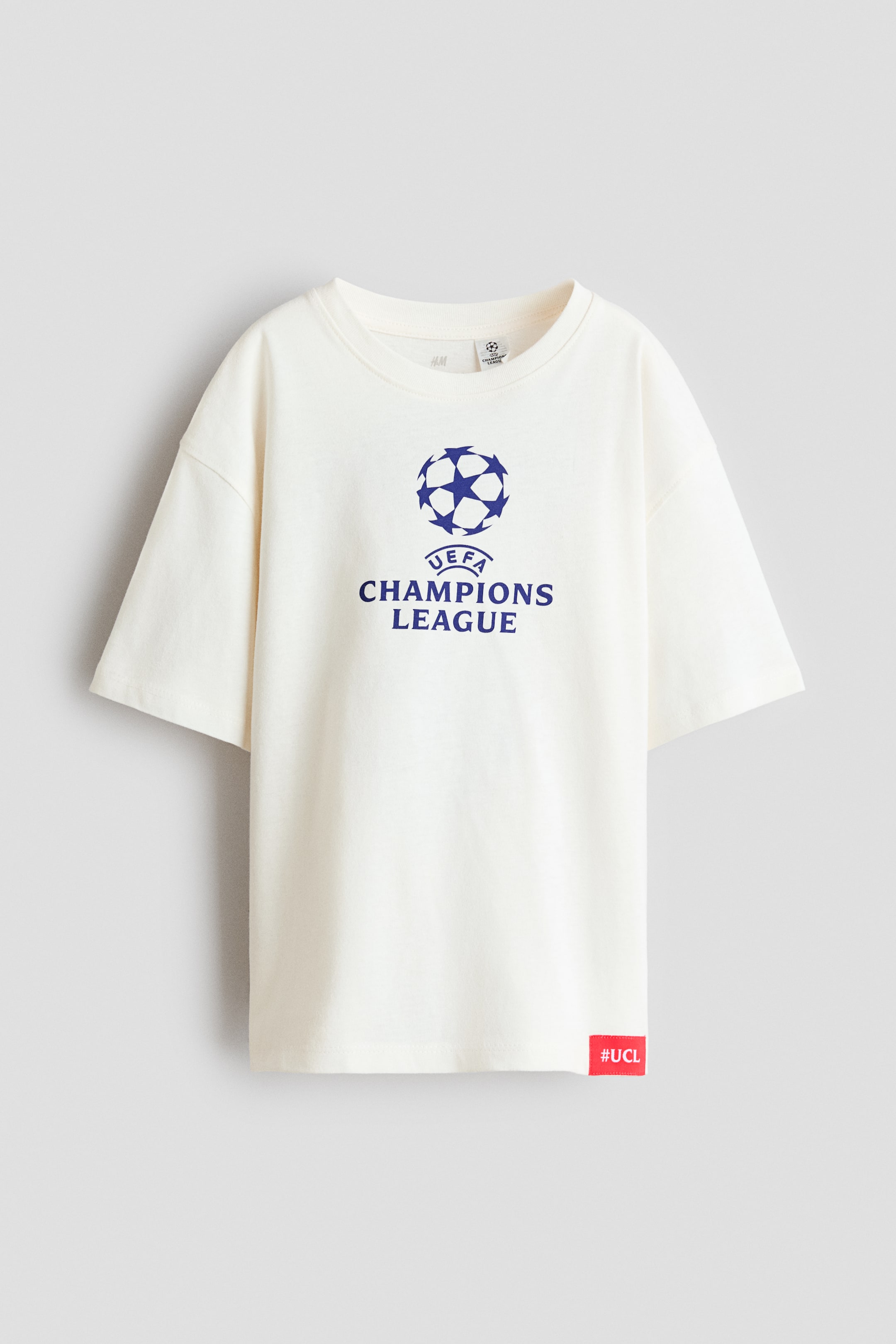 Vezi imaginea mai mare: Un tricou supradimensionat, alb murdar, este fotografiat din față, evidențiind umerii căzuți, un decolteu rotund cu margini ribate și un logo imprimat albastru „UEFA CHAMPIONS LEAGUE” pe piept. Un mic etichetă roșie cu text alb „#UCL” detaliază tivul din stânga jos.