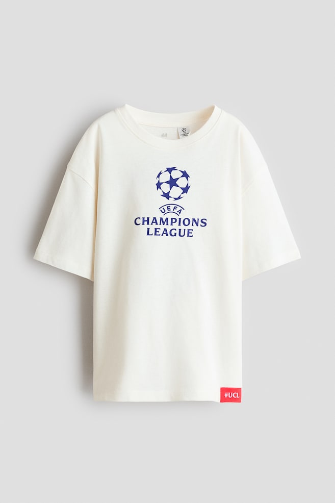 Una playera extragrande color blanco roto estÃ¡ de frente, mostrando hombros caÃ­dos, cuello redondo de ribete y un logotipo impreso azul de "UEFA CHAMPIONS LEAGUE" en el pecho. Una pequeÃ±a etiqueta roja con el texto blanco "#UCL" detalla el dobladillo inferior izquierdo. - 1