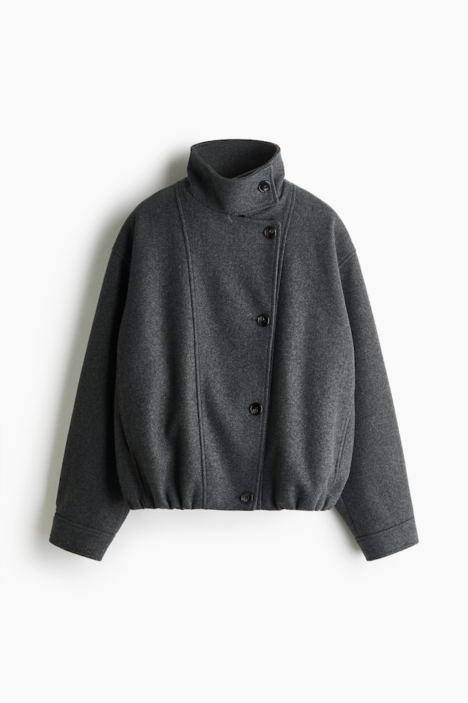 A dark grey jacket is shown from the front, featuring an asymmetric front button fastening and a high, buttoned funnel collar. The jacket showcases dropped shoulders, long sleeves with wide cuffs, and a gathered elastic hem that creates a slightly voluminous silhouette. - 1