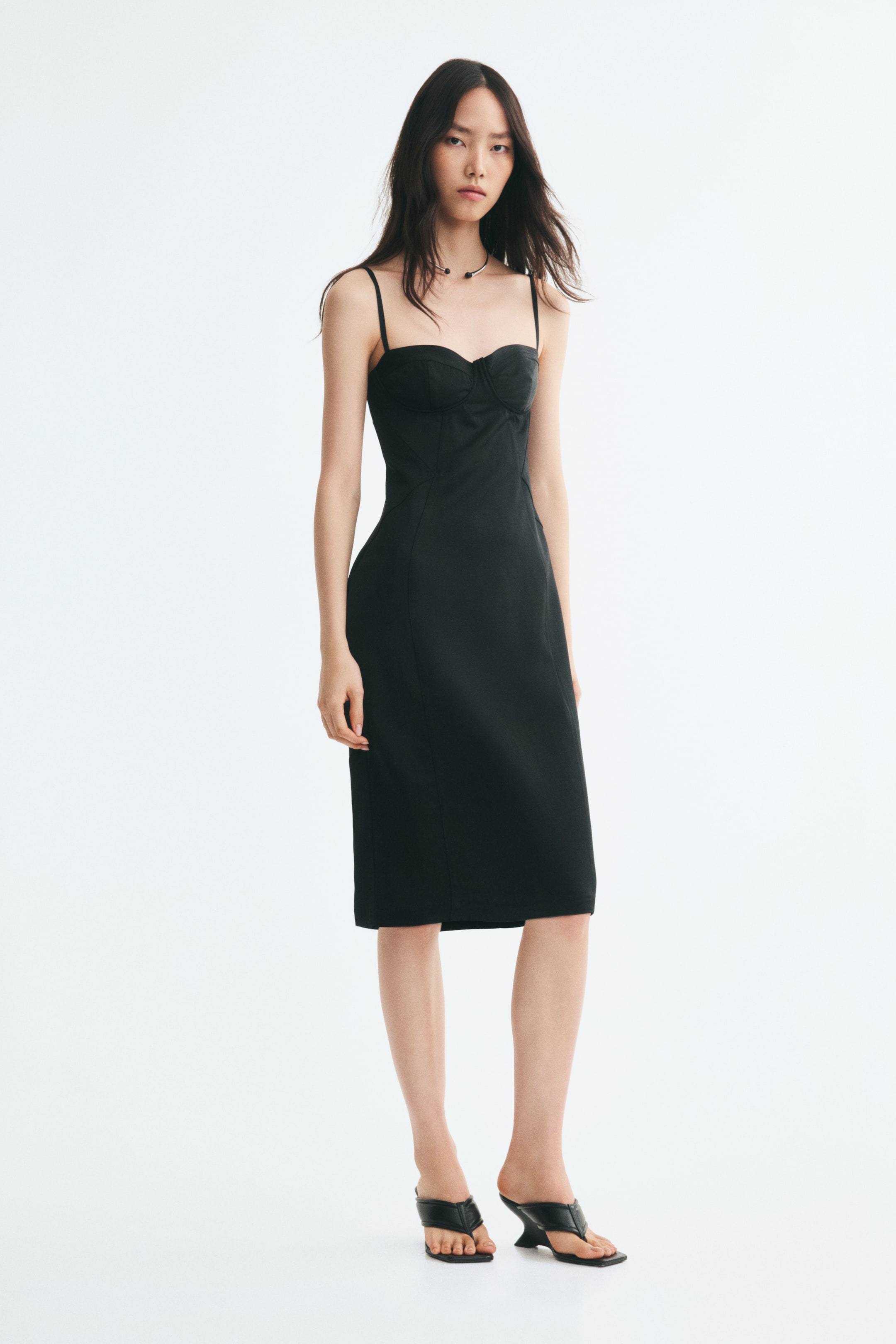 Visualizza immagine più grande: La modella indossa un abito bodycon nero lungo fino al polpaccio, caratterizzato da scollo a cuore, coppe con ferretto e sottili spalline. L’abito presenta cuciture verticali modellanti ben visibili sul davanti ed è abbinato a sandali neri con tacco.