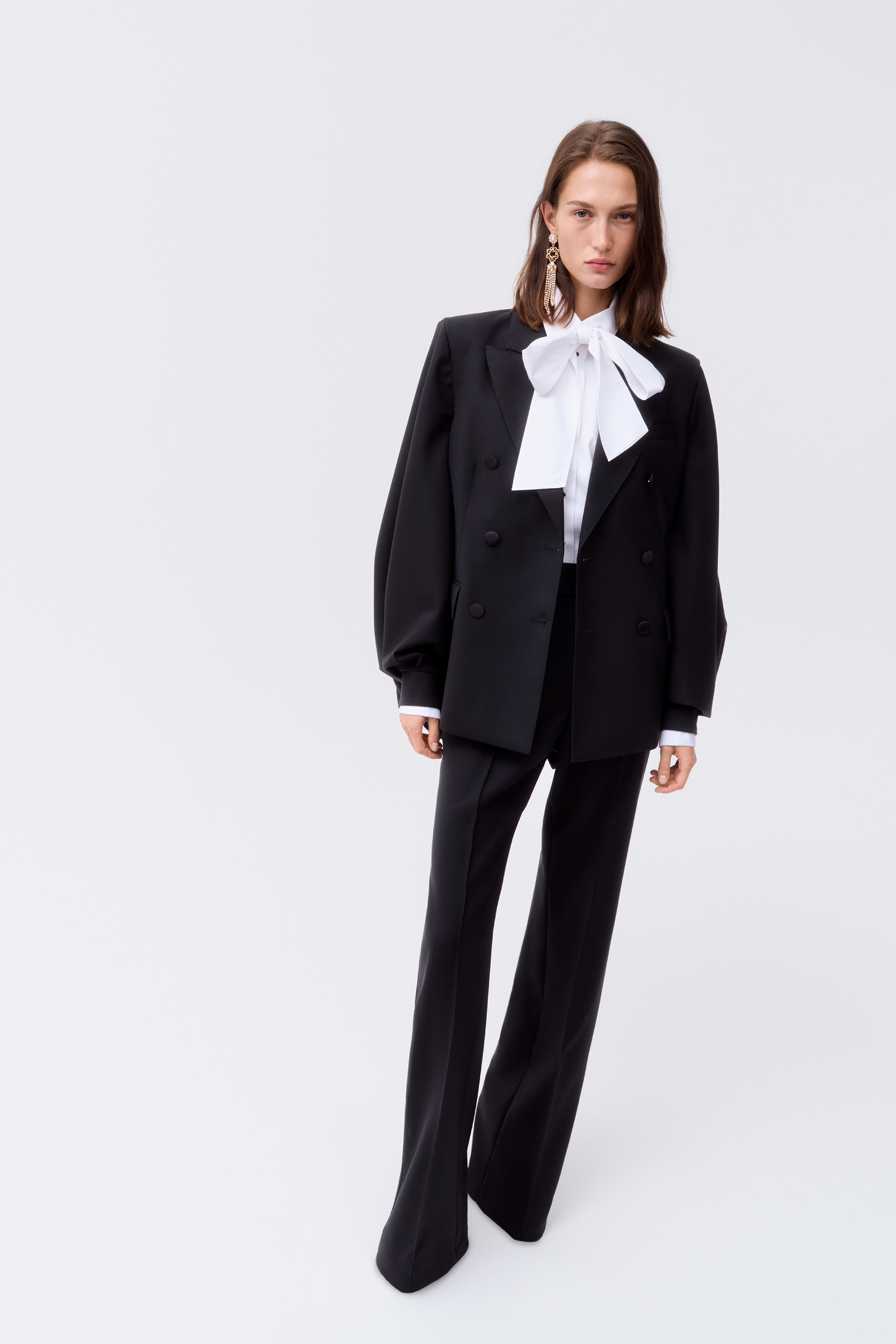 A black double-breasted wool blazer is shown facing forward, featuring structured shoulders, sharp peak lapels, and a six-button front closure. The garment has voluminous, gathered sleeves with wide cuffs and two classic flap pockets at the hips.