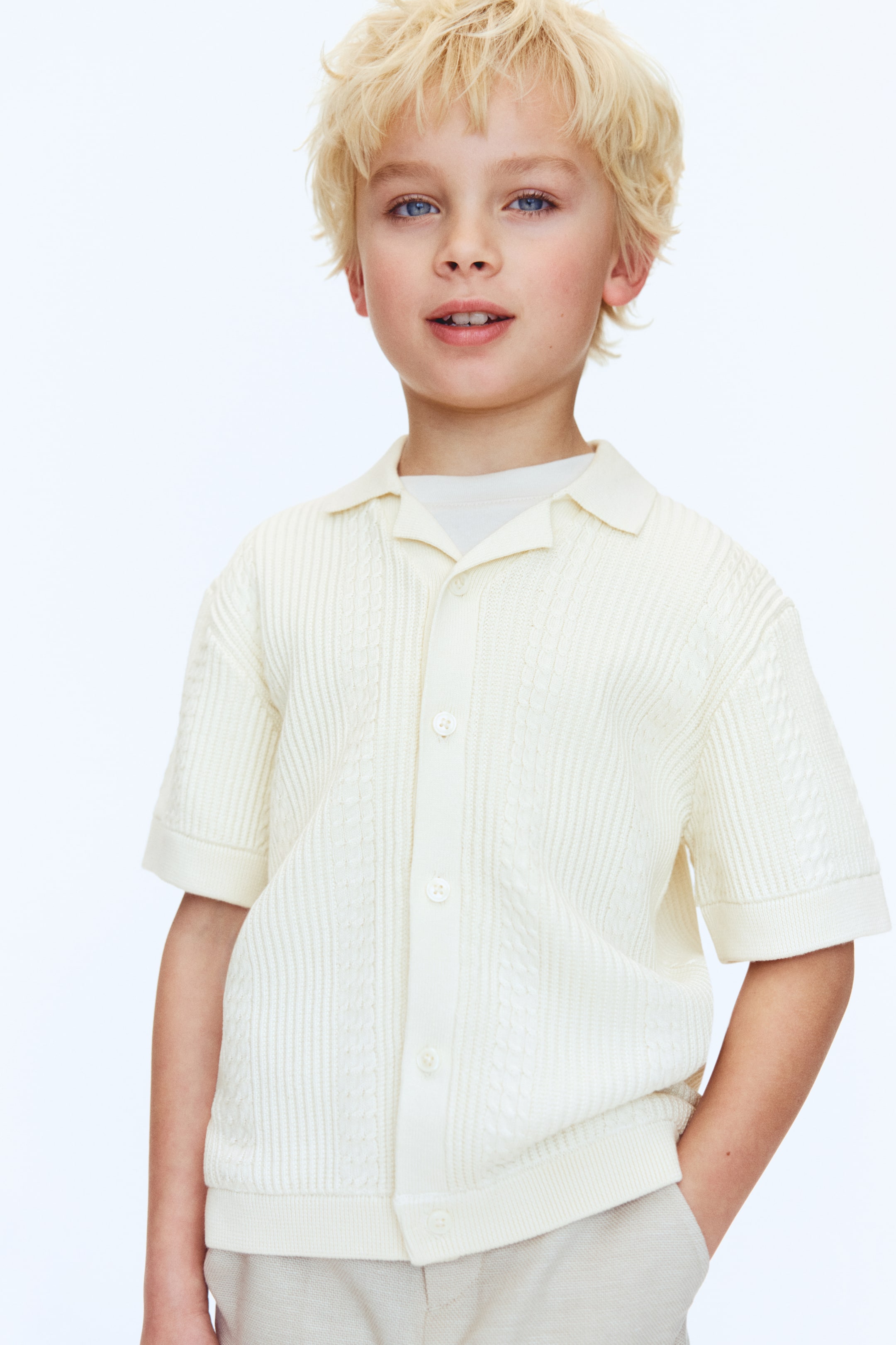 Visualizza immagine più grande: Una maglietta a maniche corte color crema in maglia viene presentata frontalmente, sovrapposta a una t-shirt bianca. Presenta una combinazione di motivi a treccia e a coste, un'abbottonatura con bottoni visibili, un colletto aperto e polsini e orlo a coste.