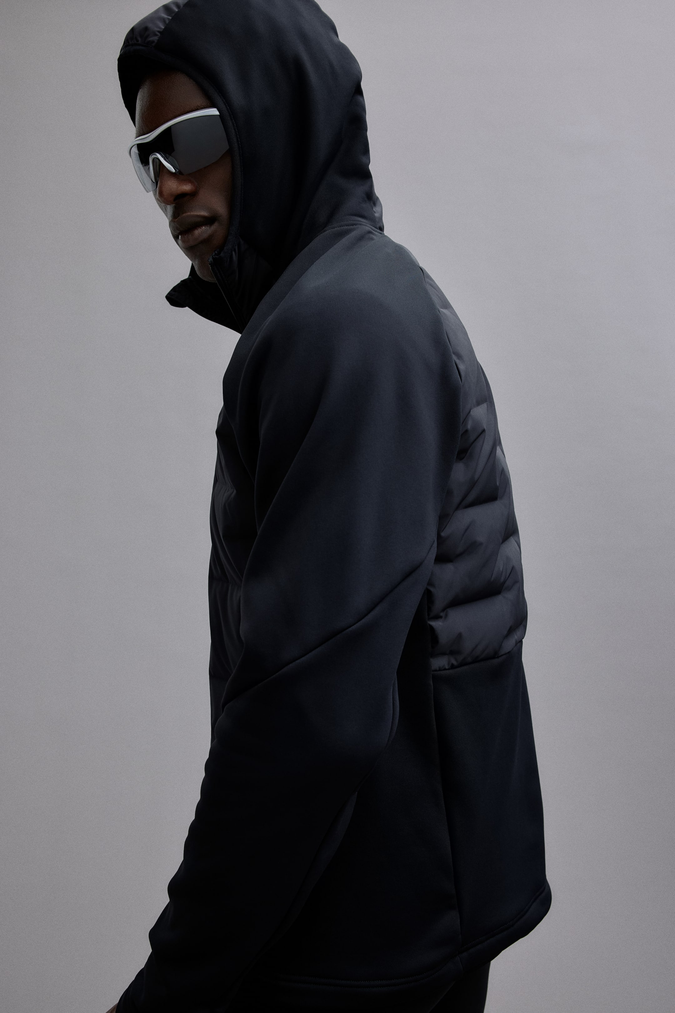 Agrandir l'image: Une veste à capuche de couleur sombre présente un tissu lisse sur les manches et la capuche. On y voit un panneau matelassé compartimenté sur le côté et le dos, vu en trois-quarts arrière.