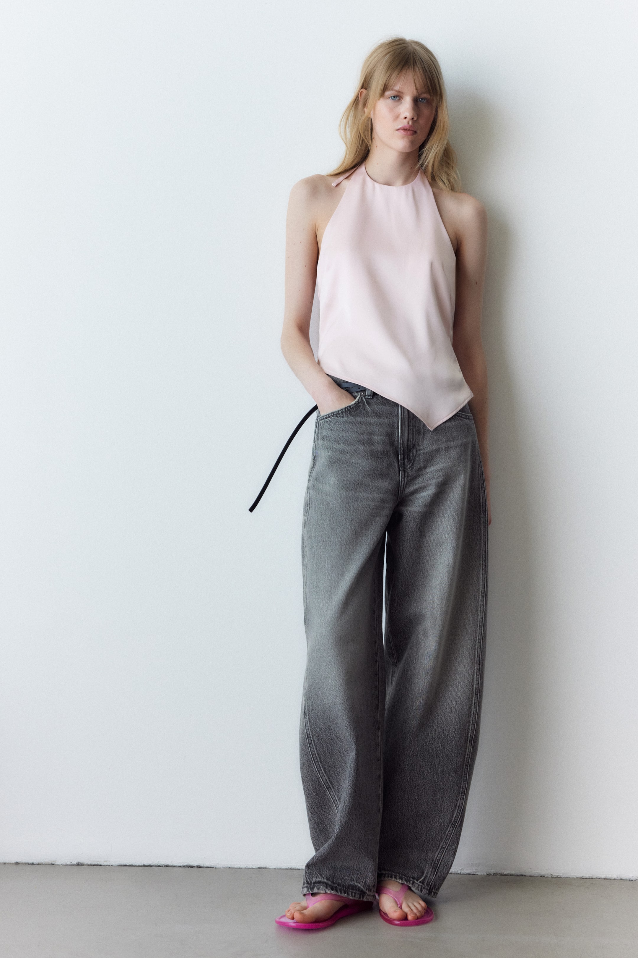 Größeres Bild anzeigen: Ein hellrosa Neckholder-Top mit asymmetrischem Saum fällt locker über ausgewaschene graue Jeans mit weitem Bein. Die Jeans bieten eine entspannte Passform, mit einem schwarzen Schnurdetail, das aus einer Vordertasche hervorragt.