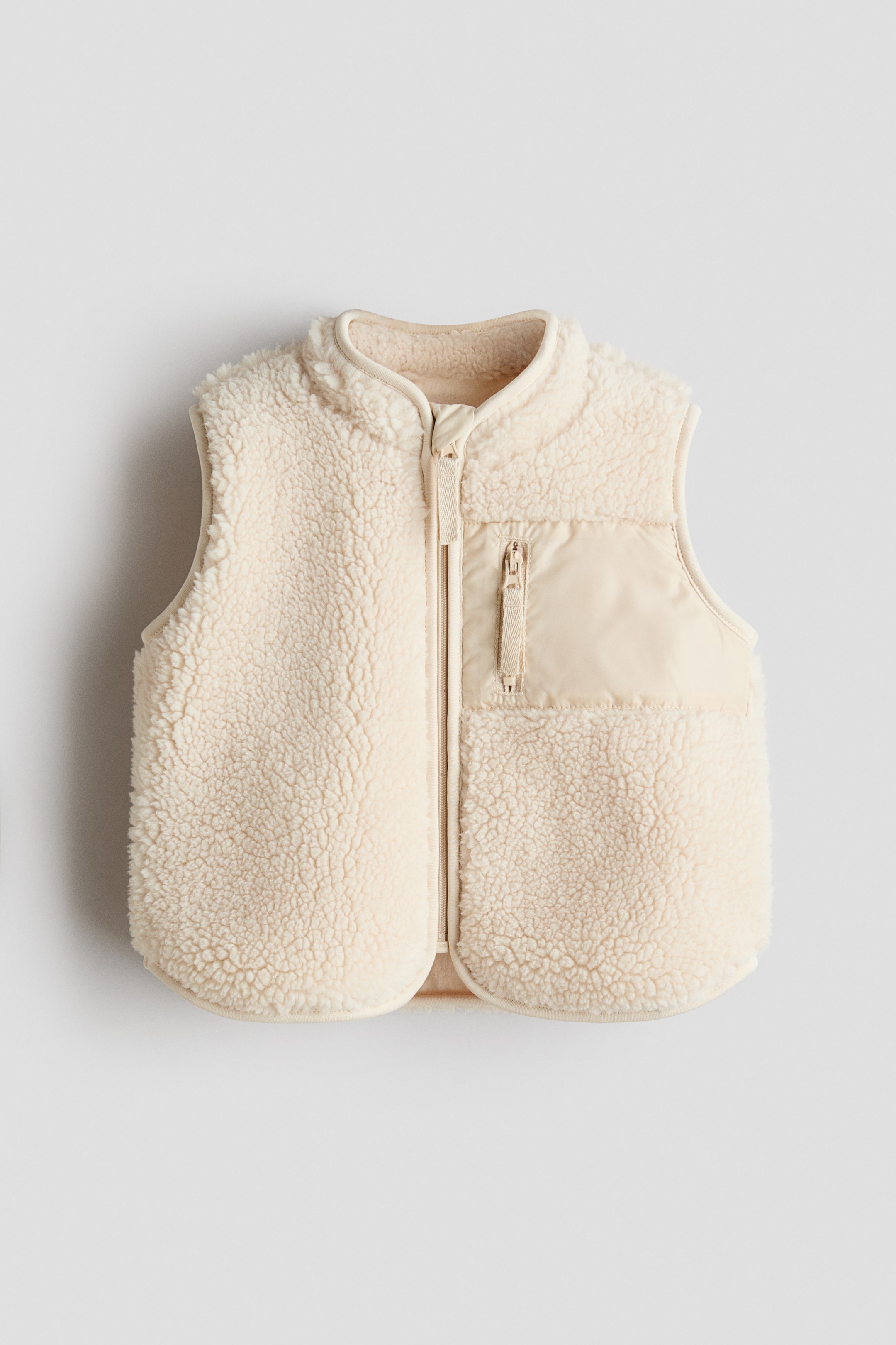 View larger image: A cream sherpa fleece vest is displayed in a front view, showcasing a full-length zip closure and a stand-up collar. A sleek, light beige zippered chest pocket is visible on the right side.