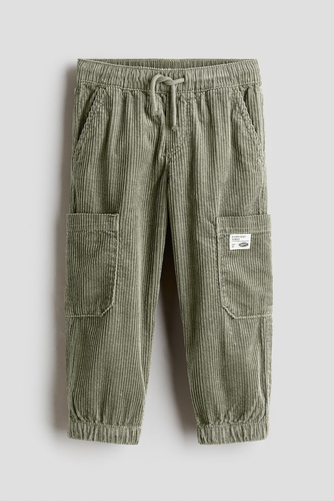A pair of sage green corduroy cargo pants is shown facing front, featuring an elasticized waistband with a matching drawstring, side pockets, and elasticized ankle cuffs. The pants are displaying large rectangular cargo pockets on the sides, with a small white patch stitched onto the left pocket. - 1