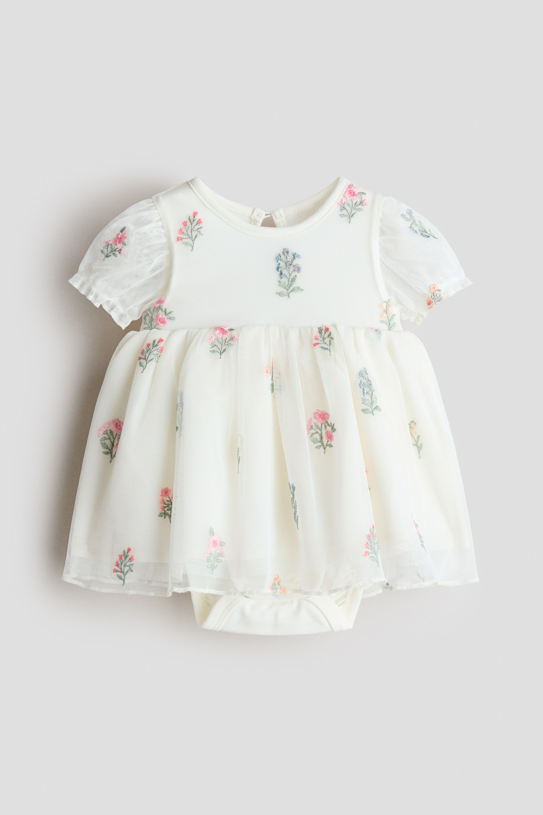 Grösseres Bild ansehen: Ein cremefarbenes Kleid mit eingenähtem Body wird von vorne präsentiert, mit einem runden Ausschnitt mit Schlüssellochöffnung und Knopf im Nacken sowie kurzen Puffärmeln mit Rüschenbesatz an den Bündchen. Eine geraffte Naht an der Taille sorgt für einen voluminösen Tüllrock, der mit bunter Blumenstickerei verziert ist, während der eingenähte Body darunter sichtbar ist und geraffte Beinnähte zeigt.