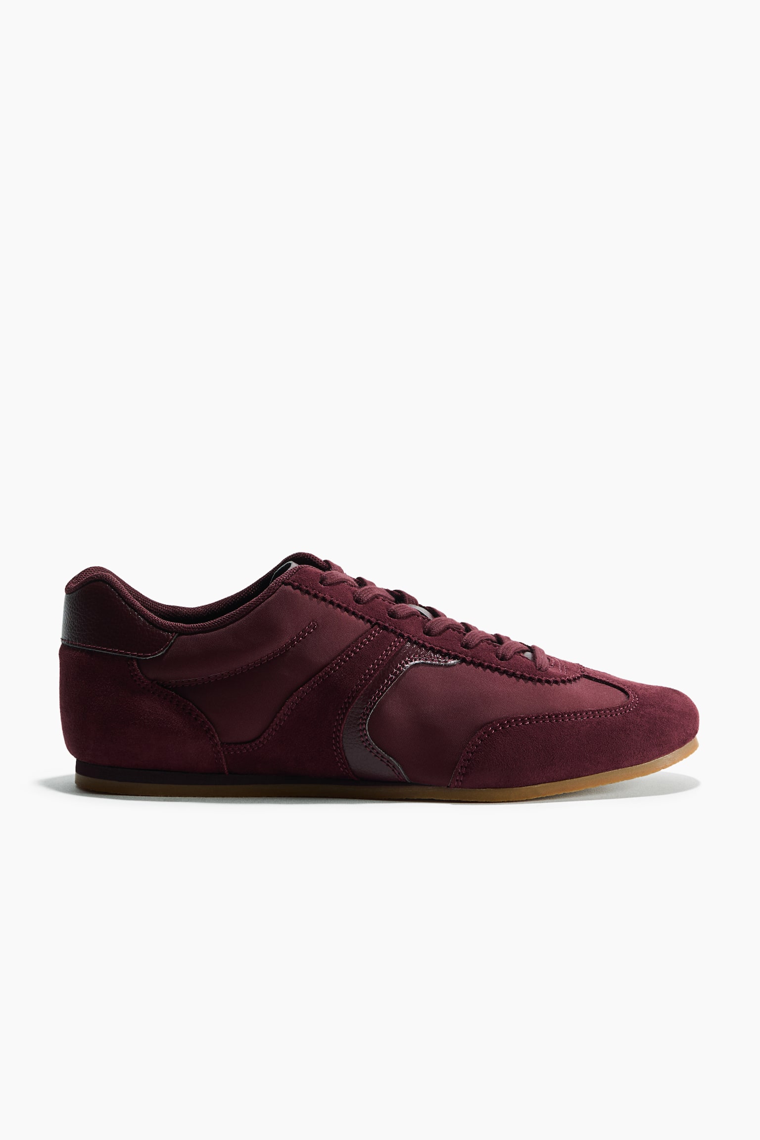 A burgundy sneaker is shown in side profile, facing right, featuring a padded top edge, tongue, and tonal lacing at the front. The shoe is crafted with mixed suede and textile panels and finished with a slim gum outsole.