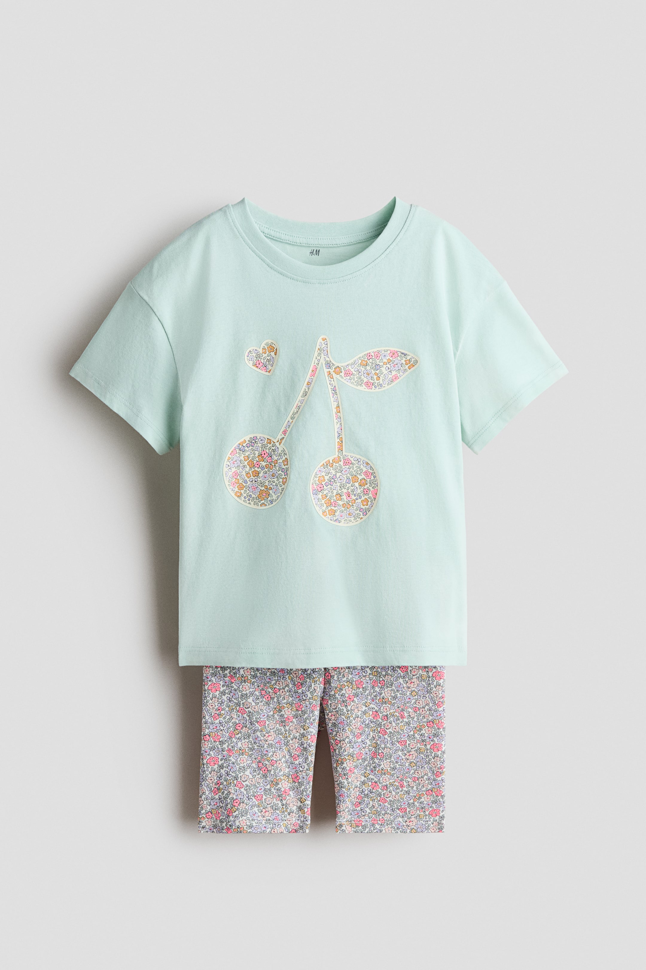 Visualizza immagine più grande: Un set composto da una t-shirt verde chiaro e pantaloncini da ciclista floreali coordinati è in mostra. La t-shirt, mostrata di FRONTE, presenta un girocollo a coste, spalle cadenti e una grande grafica di ciliegie con toppe a stampa floreale, insieme a un piccolo cuore, mentre i pantaloncini da ciclista coordinati, visibili sotto, mostrano un motivo floreale denso e una vita elasticizzata coperta.