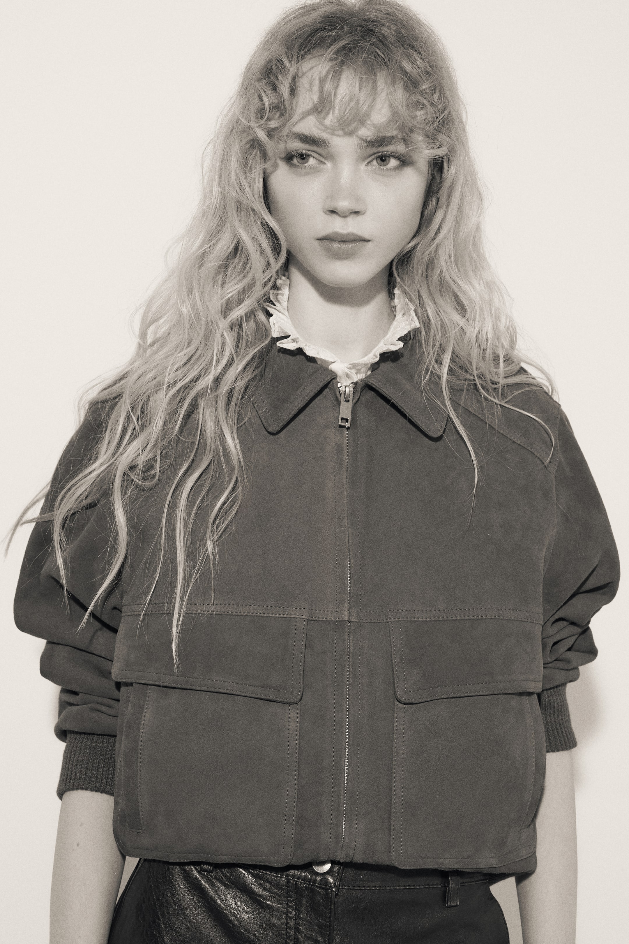 View larger image: A suede bomber jacket features a front zipper closure, a pointed collar, two square flap pockets, and long sleeves with ribbed cuffs. It is worn over a shirt with a ruffled collar and cropped to sit above black pants.