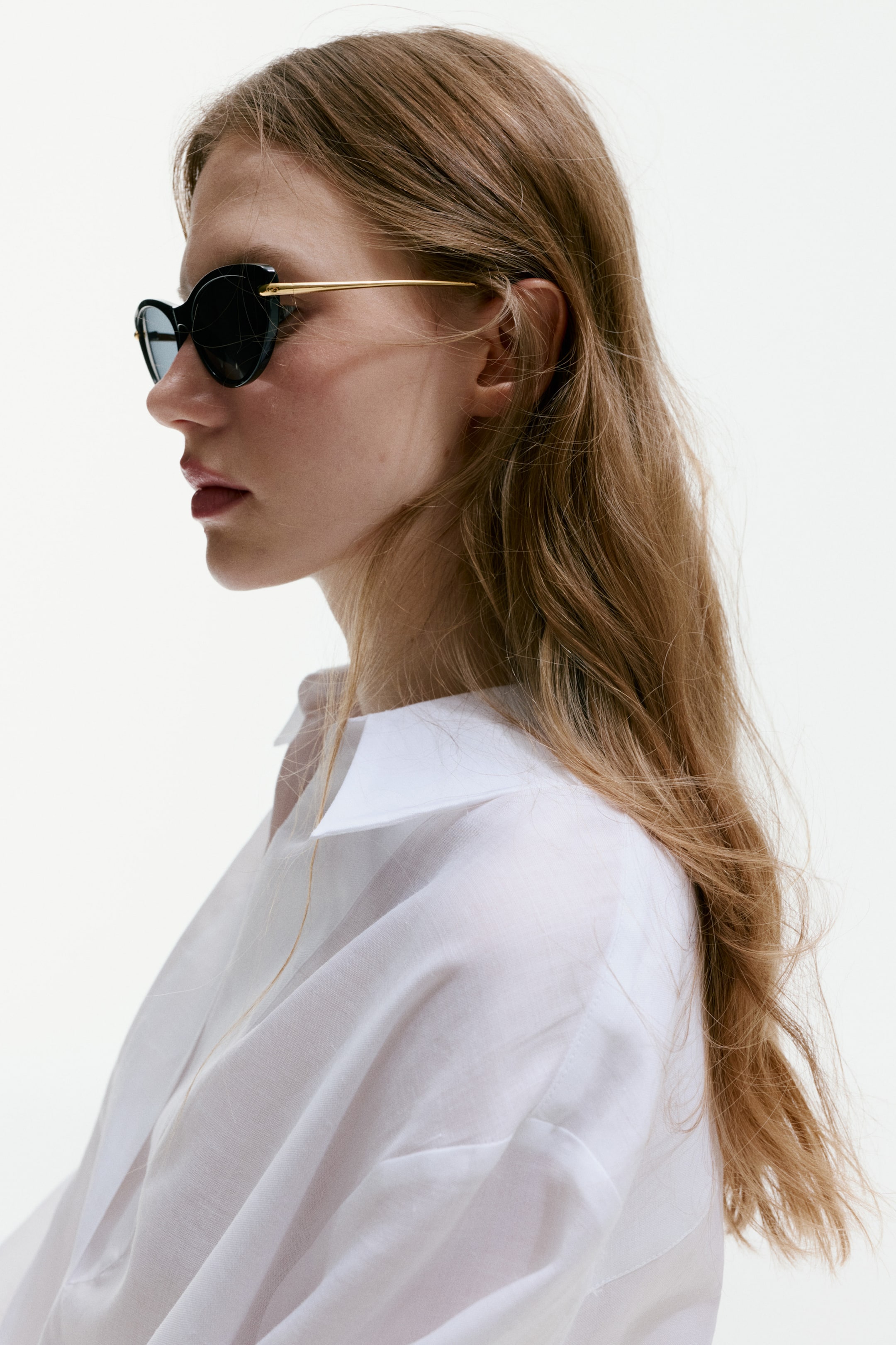 Ver imagem maior: Óculos de sol estilo "cat-eye" com armações de plástico preto. Peças laterais em metal dourado estendem-se dos cantos superiores das armações, e lentes de plástico escurecidas com proteção UV completam o design.