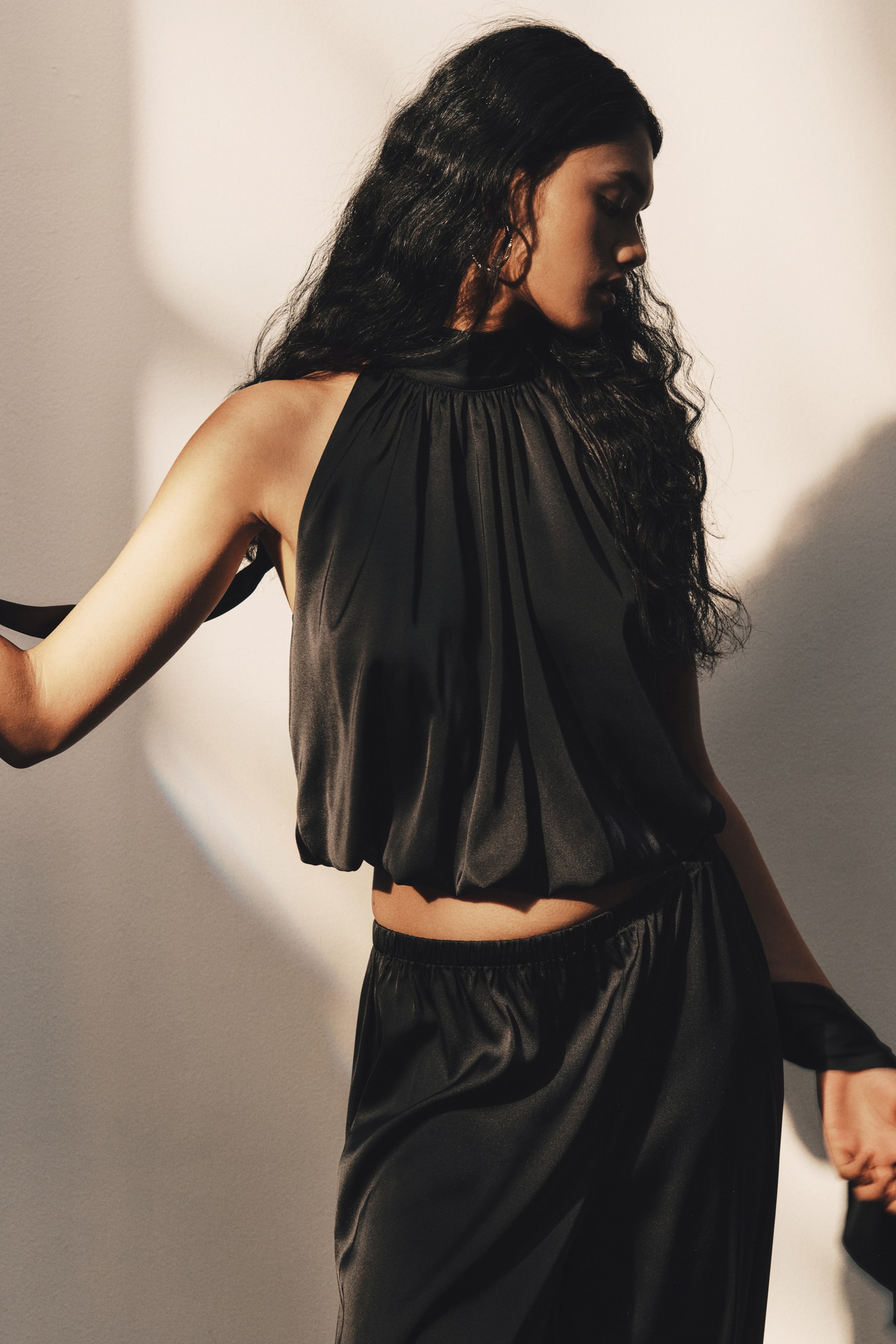 View larger image: The model is wearing a black satin halter top with a high, gathered neckline and a puffy, elasticized bubble hem. The loose-fit, sleeveless top is styled with matching black satin trousers, with long ties extending from the back of the neck.