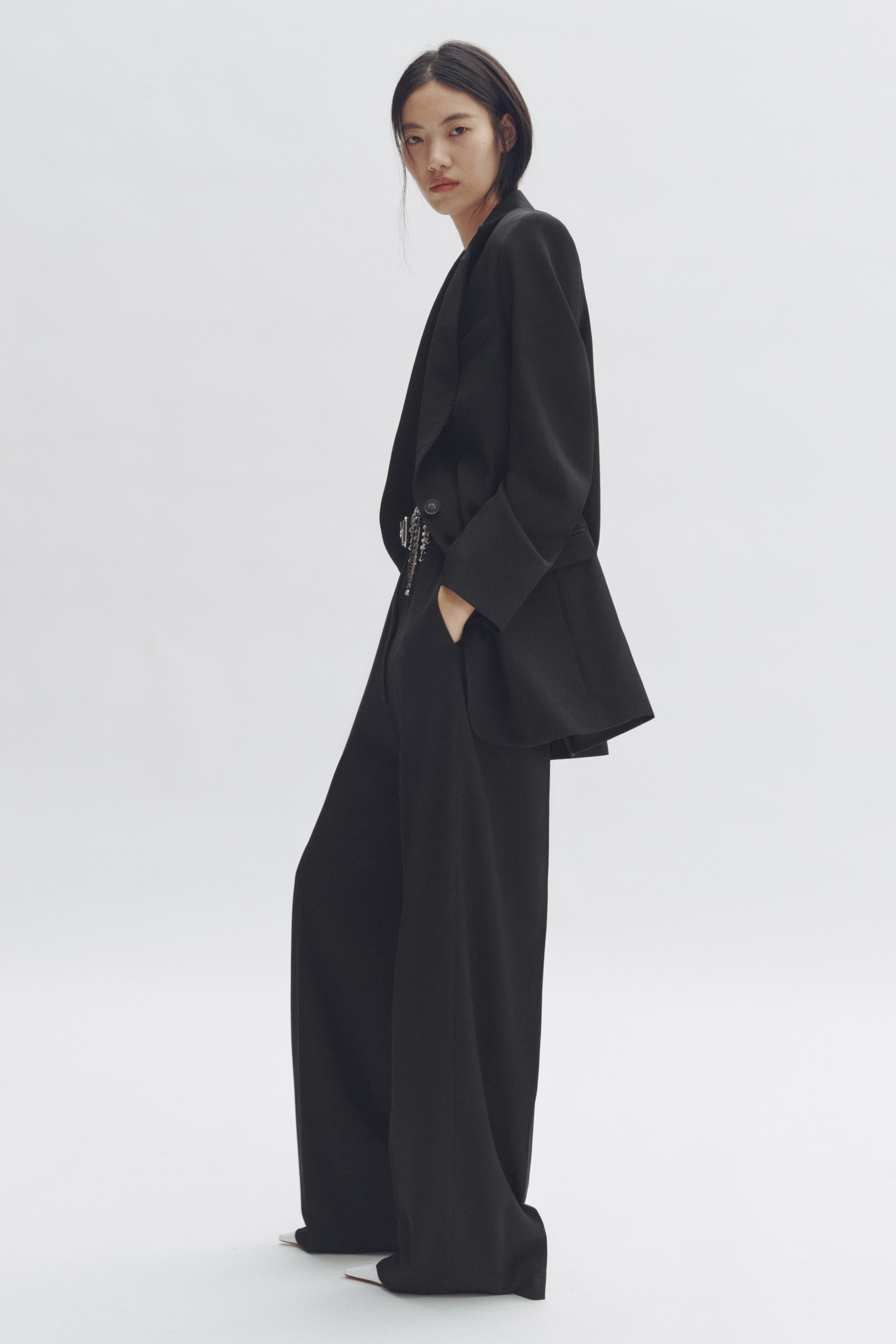 Visualizza l'immagine ingrandita: Un tailleur pantalone nero viene presentato da un'angolazione leggermente laterale, caratterizzato da un blazer oversize e pantaloni a gamba larga. Il blazer presenta ampi revers, polsini piegati e una chiusura a un bottone con un dettaglio a catena argentata, mentre i pantaloni mostrano una vestibilità lunga e fluente.