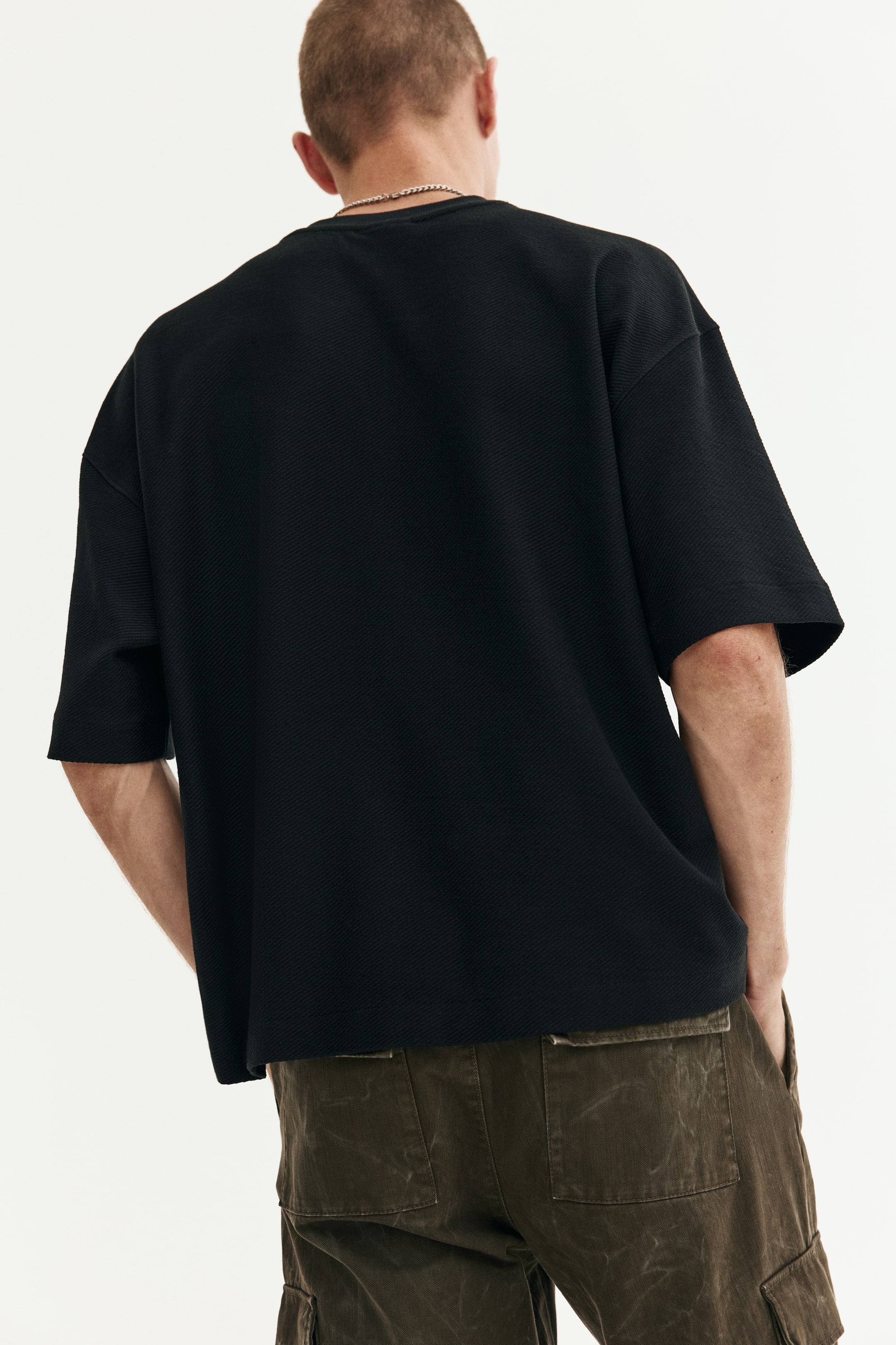 View larger image: A black t-shirt is being shown from the back, featuring an oversized, relaxed fit with wide short sleeves. The fabric showcases a subtle diagonal ribbed knit texture throughout.