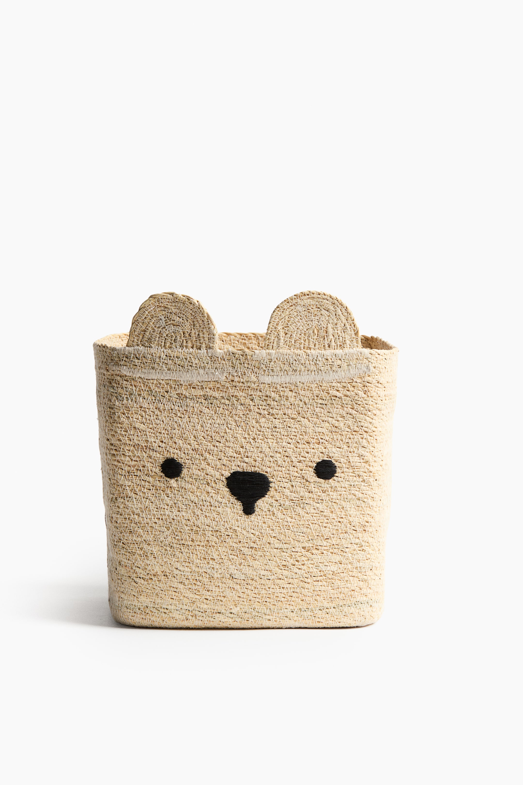View larger image: A bear-shaped straw storage basket is facing forward, displaying a natural beige woven texture with two rounded ears extending from the top rim. The front of the basket features embroidered black eyes and a nose to form a bear's face.
