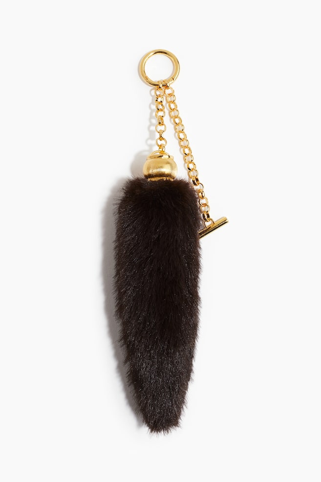 A dark brown, tapered fur keychain is hanging vertically. It features a ridged gold-tone cap, from which a gold-tone chain with a small T-bar detail is extending upwards to a large O-ring. - 1