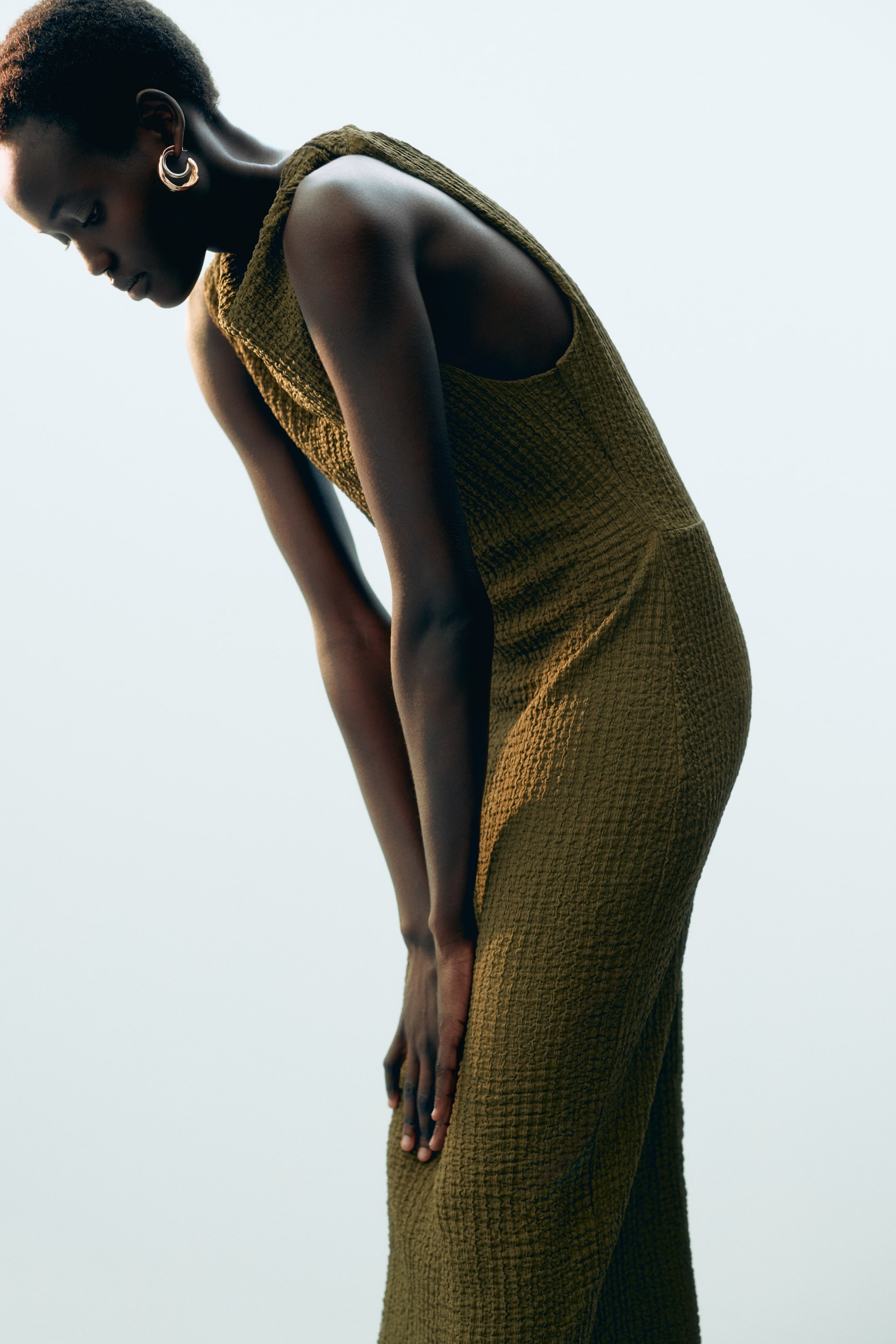 View larger image: The model is wearing a fitted midi dress in olive textured jersey, showcasing an asymmetric neckline and a twisted shoulder strap that creates a softly draped effect. She is leaning forward with her hands resting on her knees.