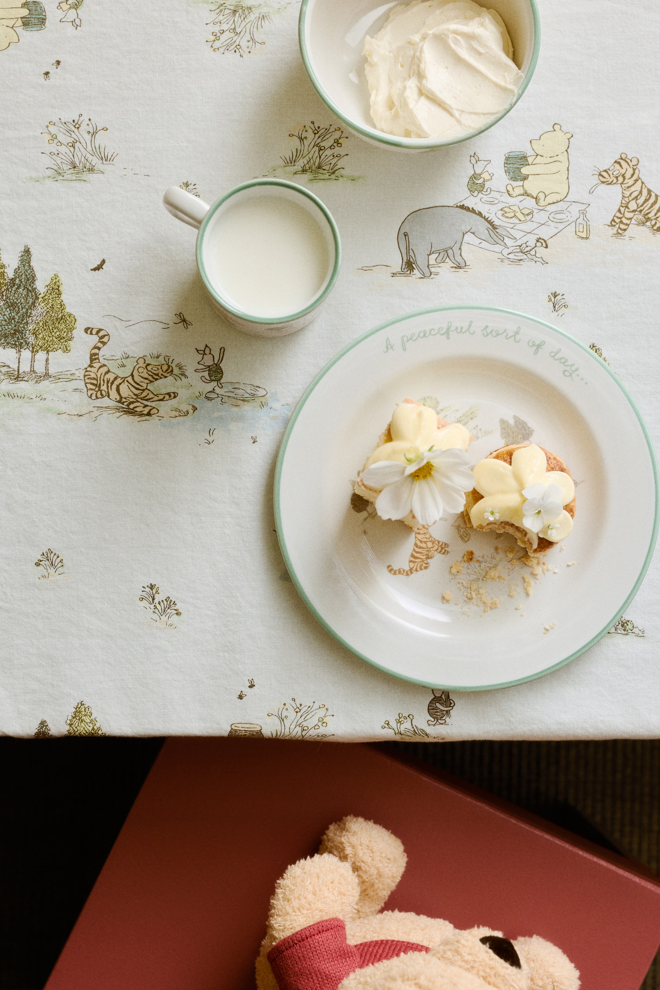 View larger image: A tablecloth featuring a light-coloured fabric with a scattered print of Winnie the Pooh characters and natural elements. The illustrations display a muted, line-drawing style in brown, green, and yellow tones.