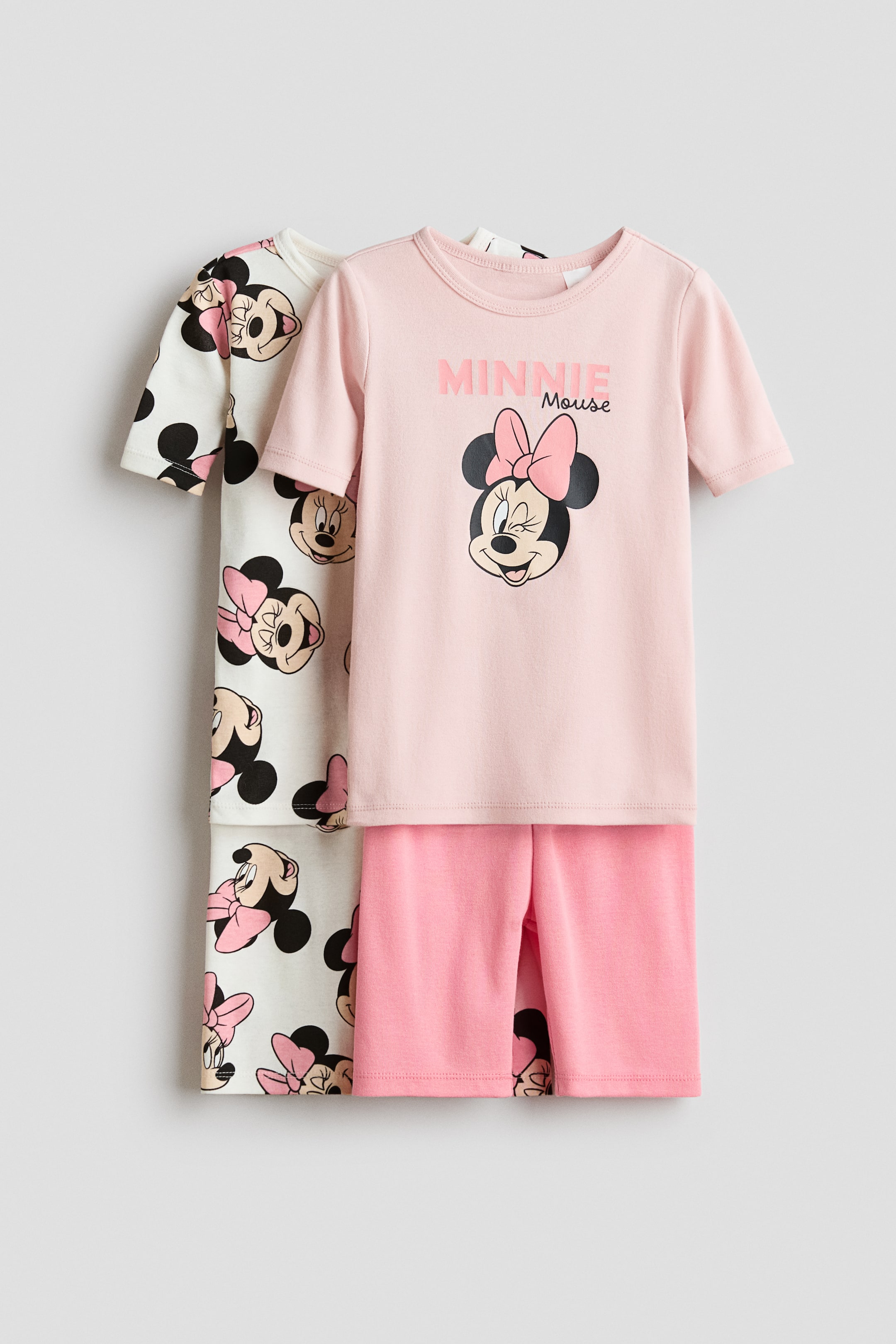 Voir une image plus grande: Deux ensembles de pyjamas sont présentés, avec le premier ensemble mettant en vedette un t-shirt à manches courtes rose pâle arborant un motif de Minnie Mouse et des shorts assortis. Un deuxième ensemble, partiellement visible à l'arrière, montre un t-shirt crème à manches courtes et des shorts avec un motif intégral de Minnie Mouse et de pois.