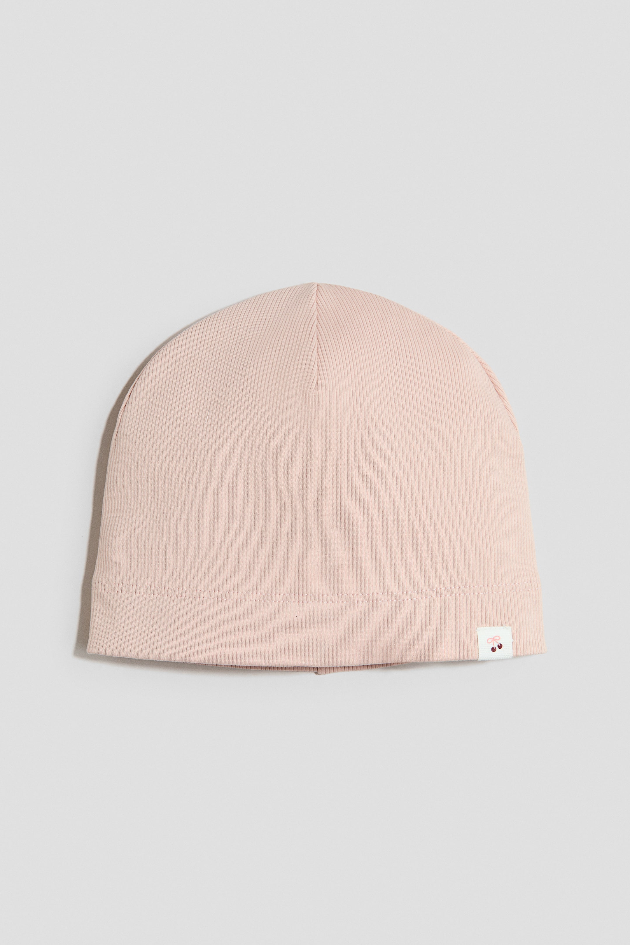 View larger image: A pale pink ribbed cotton-jersey beanie lies flat, front-facing, with a small white fabric label bearing a red cherry motif on the right front of its folded hem.
