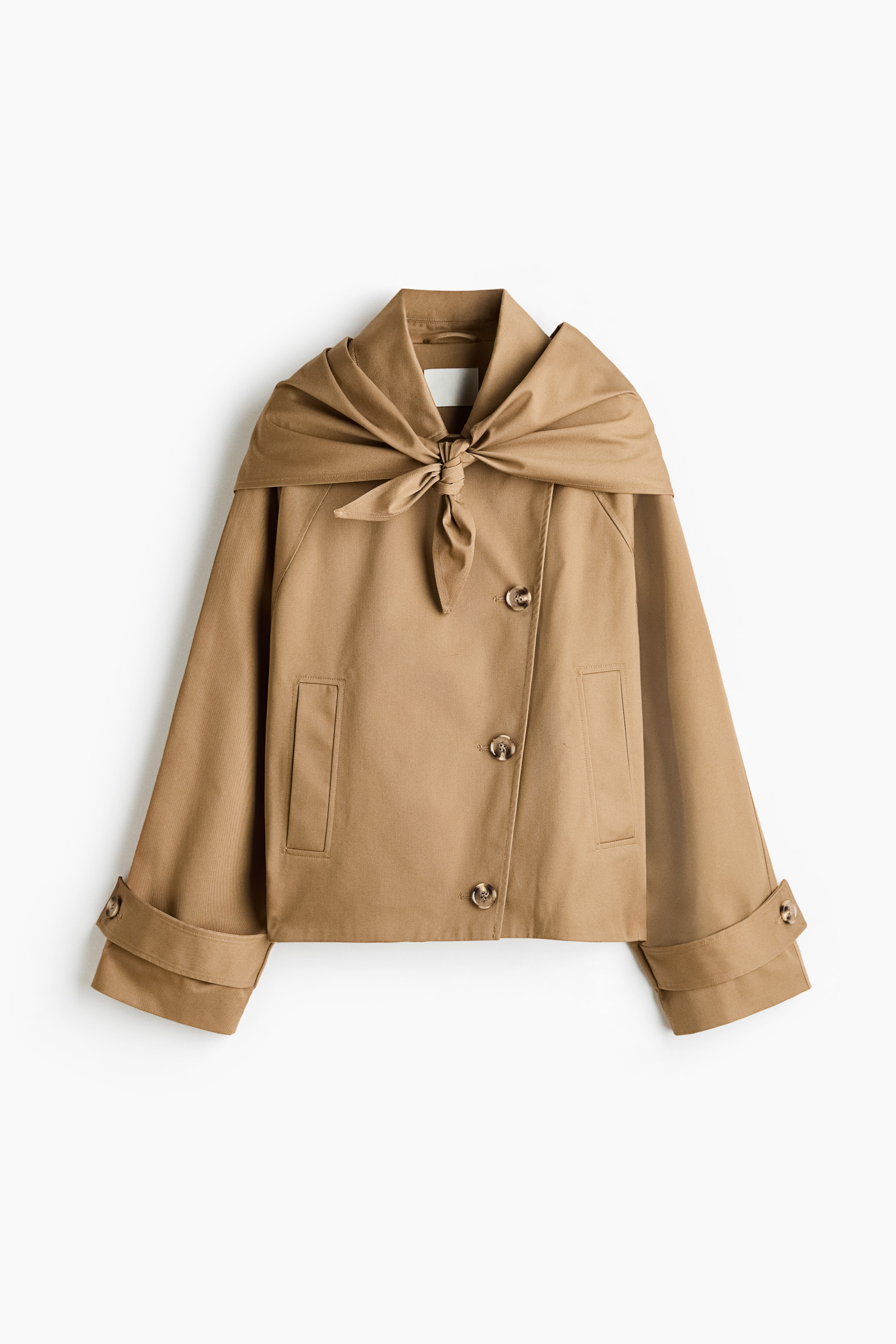 Ingrandisci l'immagine: Un corto cappotto trench oversize in twill color cammello mostra il suo davanti, esibendo un colletto, una chiusura monopetto sfalsata con tre bottoni e due tasche anteriori a filetto. Le sue maniche a pipistrello, con taglio raglan, presentano linguette e bottoni ai polsini, mentre una sciarpa triangolare coordinata è annodata sul davanti.