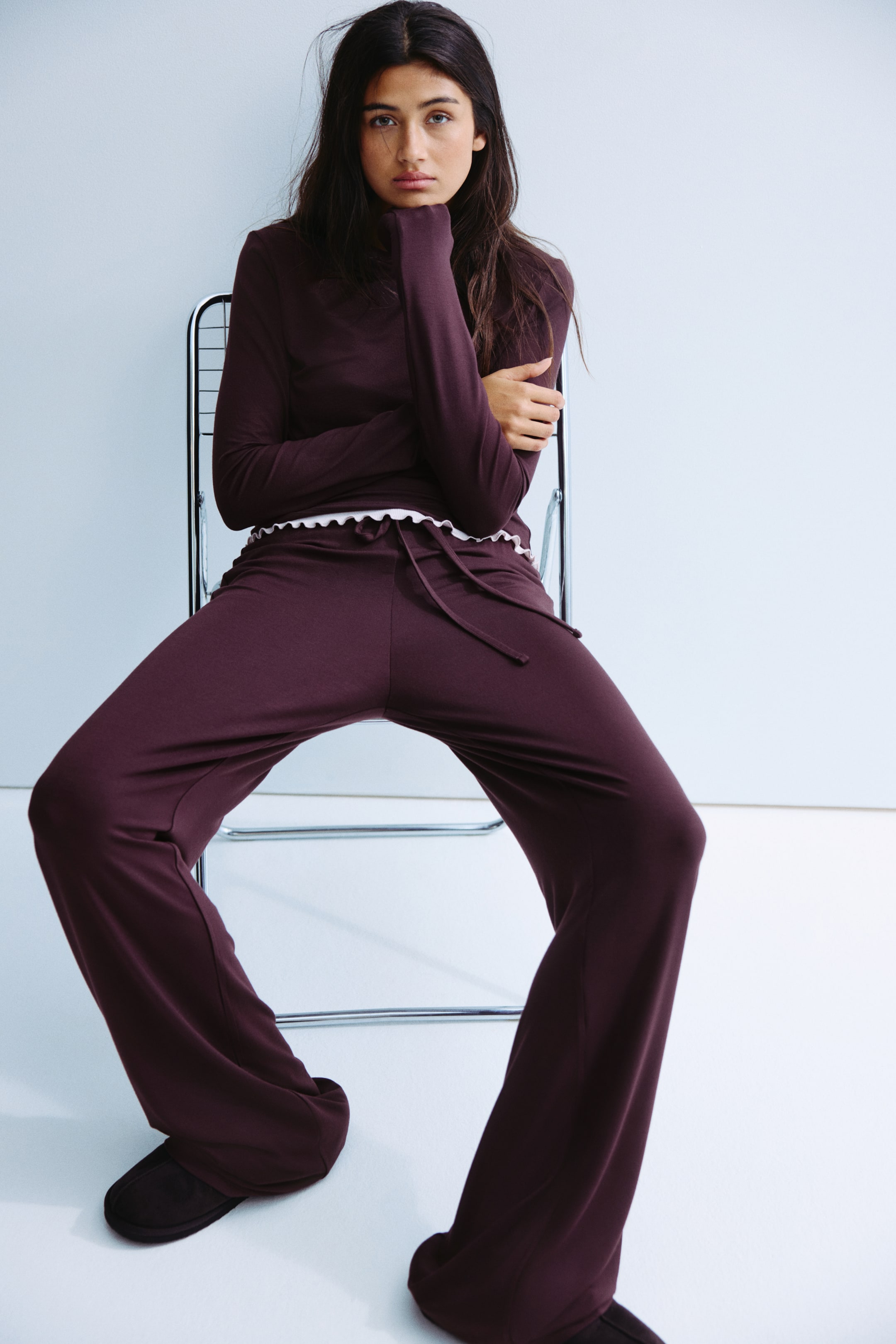 Grotere afbeelding bekijken: Een loungewear set in donkere pruimen kleur, bestaande uit een top met lange mouwen en hoge hals, en broeken met wijde pijpen, wordt gepresenteerd in vooraanzicht. De top heeft een wit geribbeld randje langs de zoom en de broeken hebben een tailleband met trekkoord.