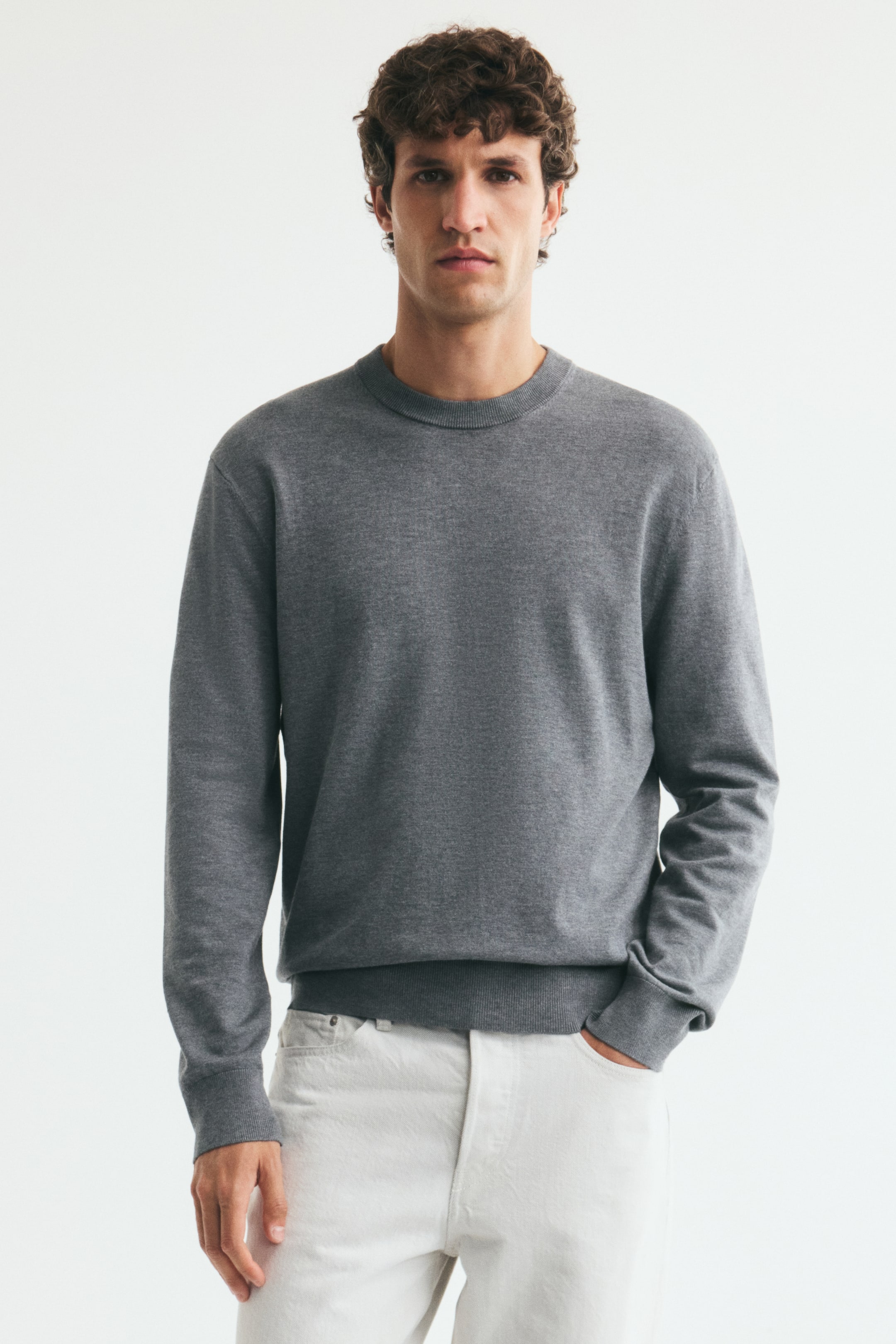 Agrandir l'image: Un pull fin en maille grise est porté par le mannequin, avec une coupe regular, un col rond côtelé et des bords-côtes aux poignets et à l'ourlet. Le pull est associé à un jean blanc, le mannequin glissant une main dans la poche avant.