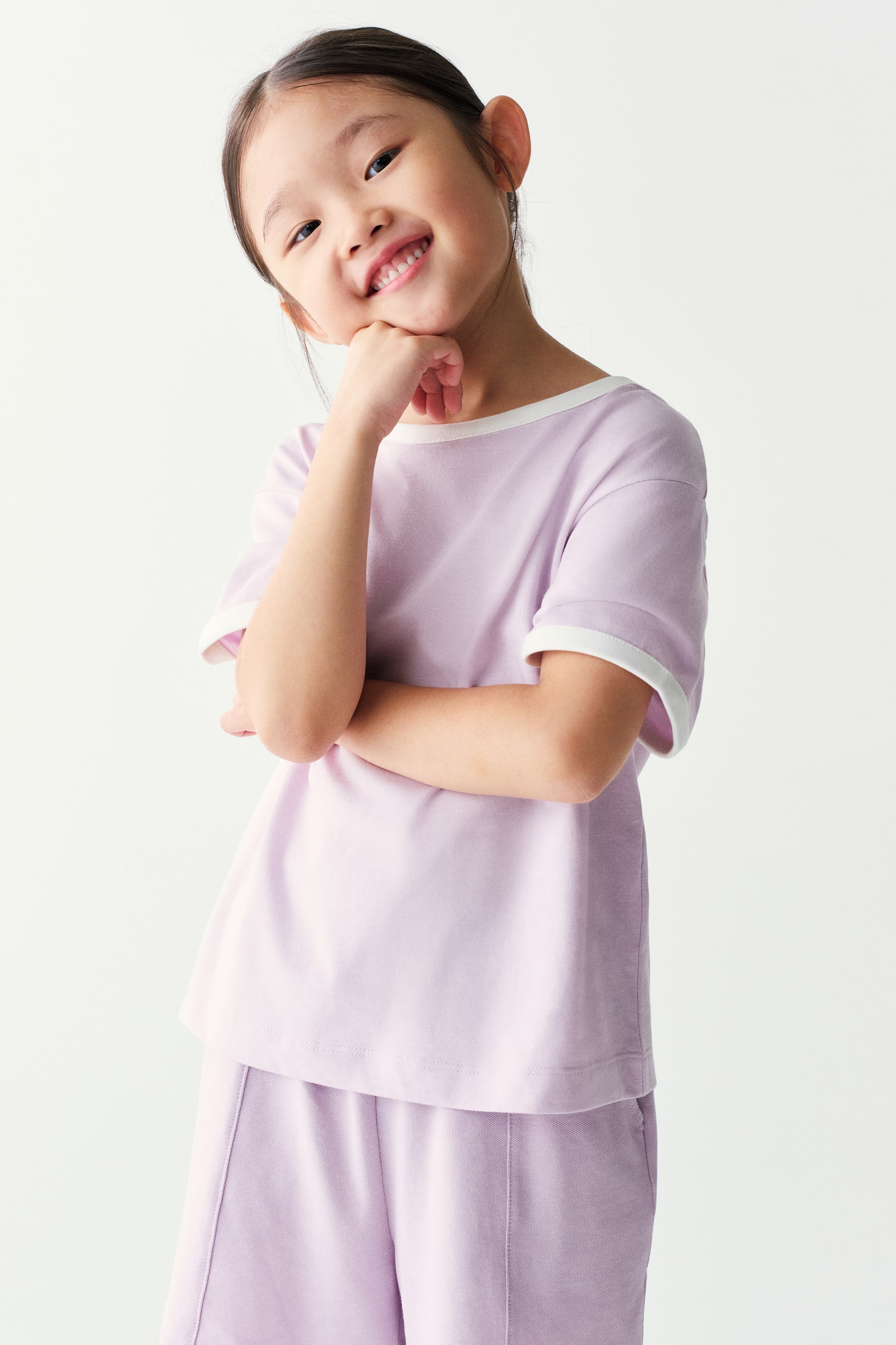 View larger image: A light purple, short-sleeved ringer t-shirt with contrasting white trim on the crew neck and cuffs is worn by the model, paired with matching light purple shorts. The model is posing with crossed arms while resting their chin on their hand.